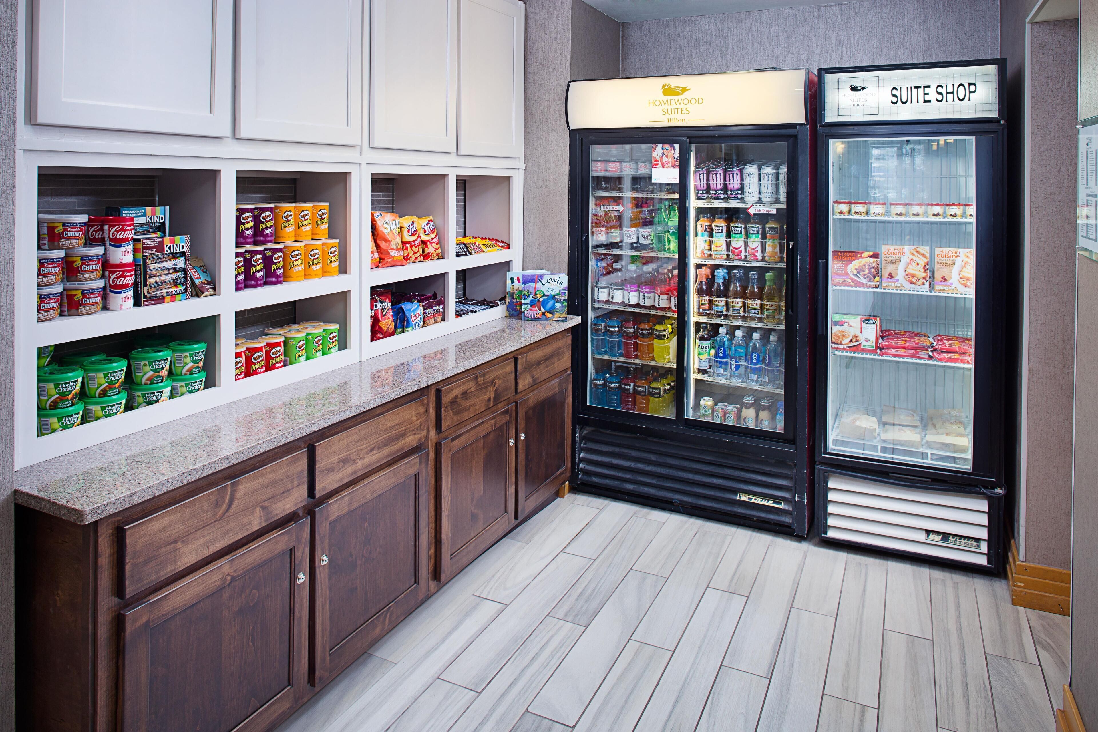 A kitchenette with a refrigerator stocked with beverages and snacks, a shelf with packaged food items, and wooden cabinets.