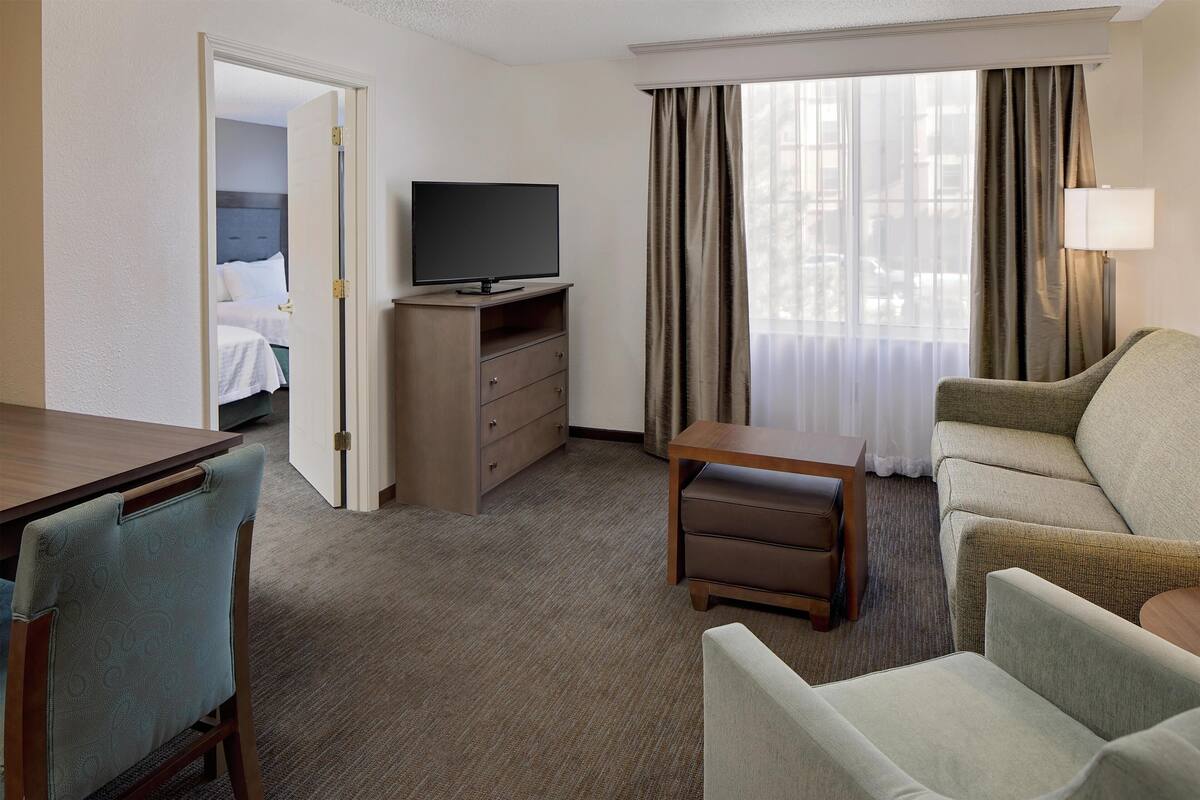 A hotel room with a bed, a TV on a stand, a desk, a chair, a sofa, and a window with curtains.