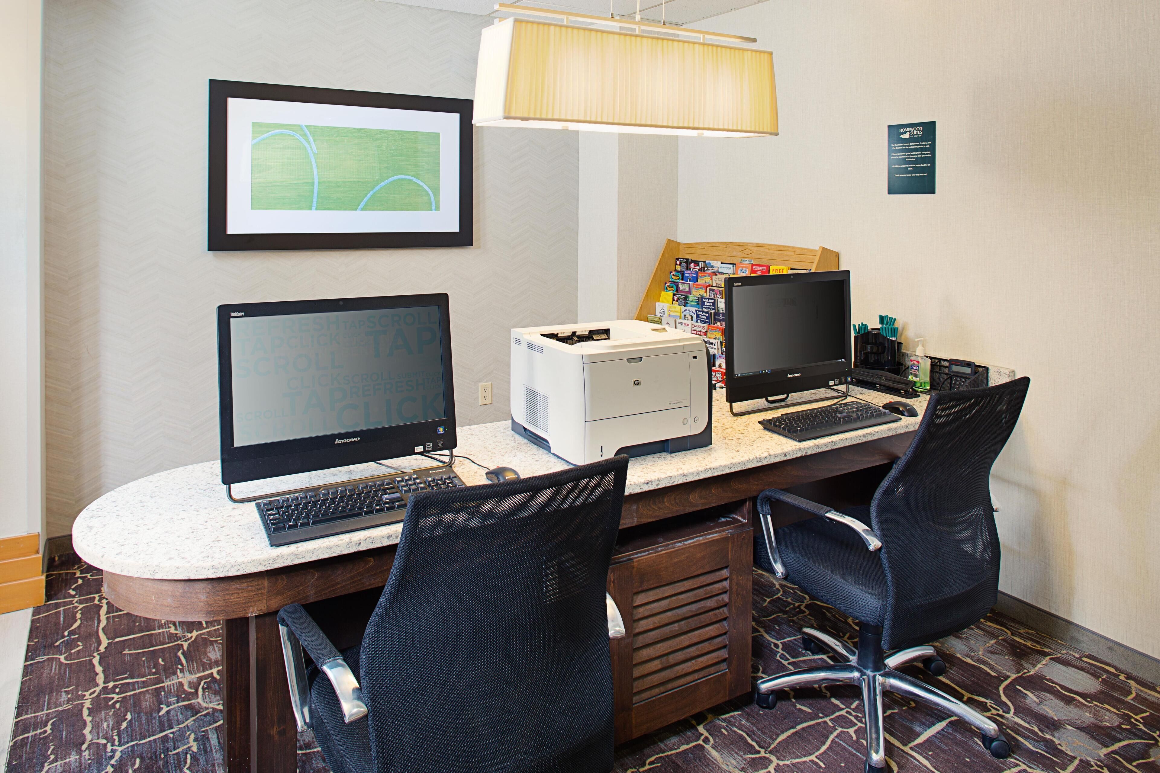 Um espaço de trabalho em hotel com dois monitores, uma impressora e uma lâmpada de mesa.