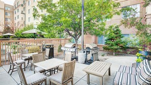 An outdoor patio area with wooden tables and chairs, a barbecue grill, and a large tree providing shade. - Homewood Suites by Hilton Albuquerque Uptown (Albuquerque)