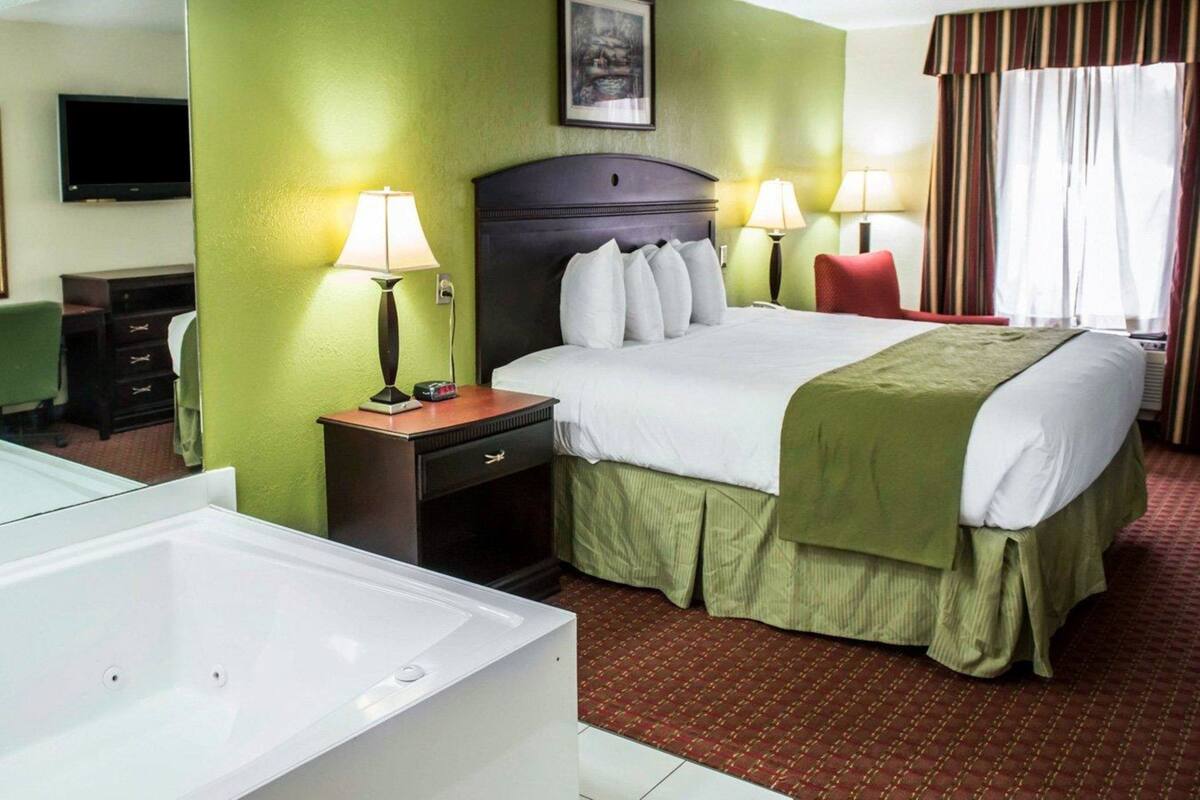 A hotel room with a large bed, two bedside lamps, a flat-screen TV, a desk with a chair, and a large white bathtub.