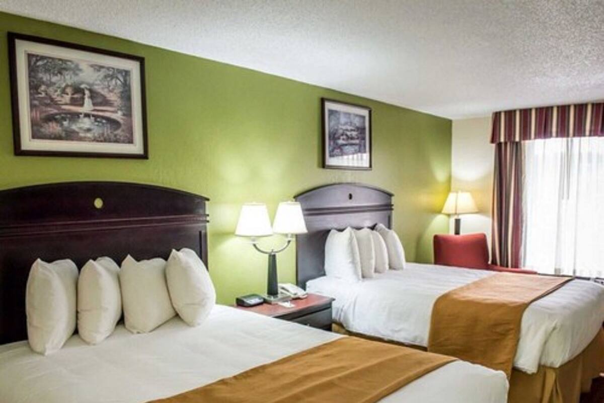 A hotel room with two beds, green walls, framed pictures, bedside lamps, and a window with curtains.