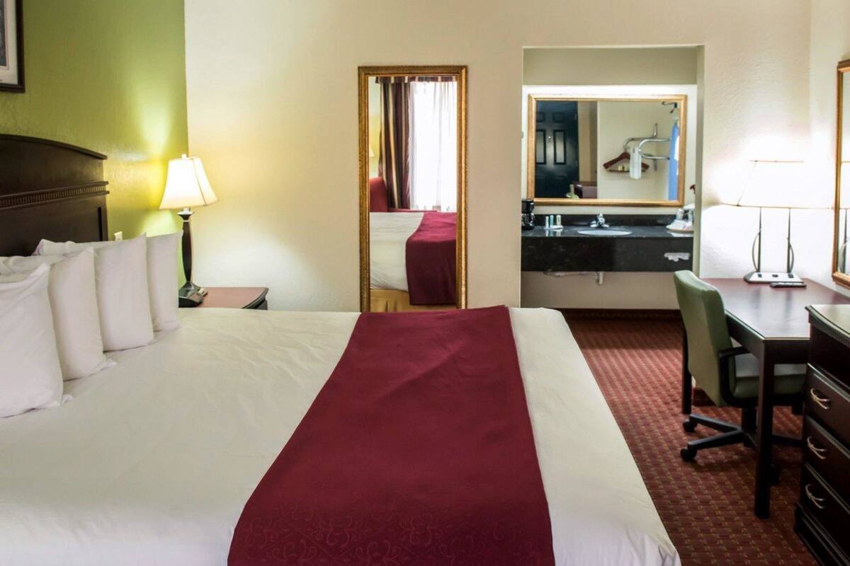 A hotel room with a large bed, a desk, a chair, a mirror, and a bathroom visible through an open door.
