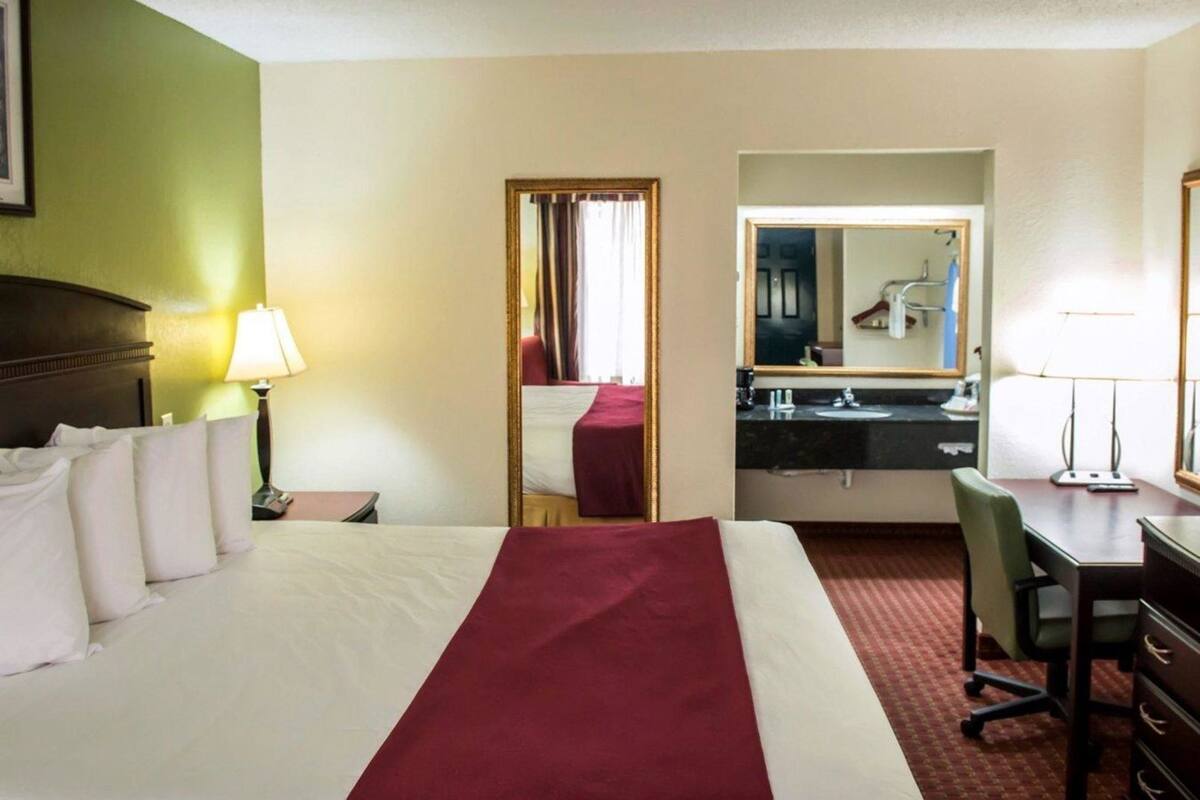A hotel room with a bed, a desk, a chair, a mirror, and a bathroom visible through an open door.