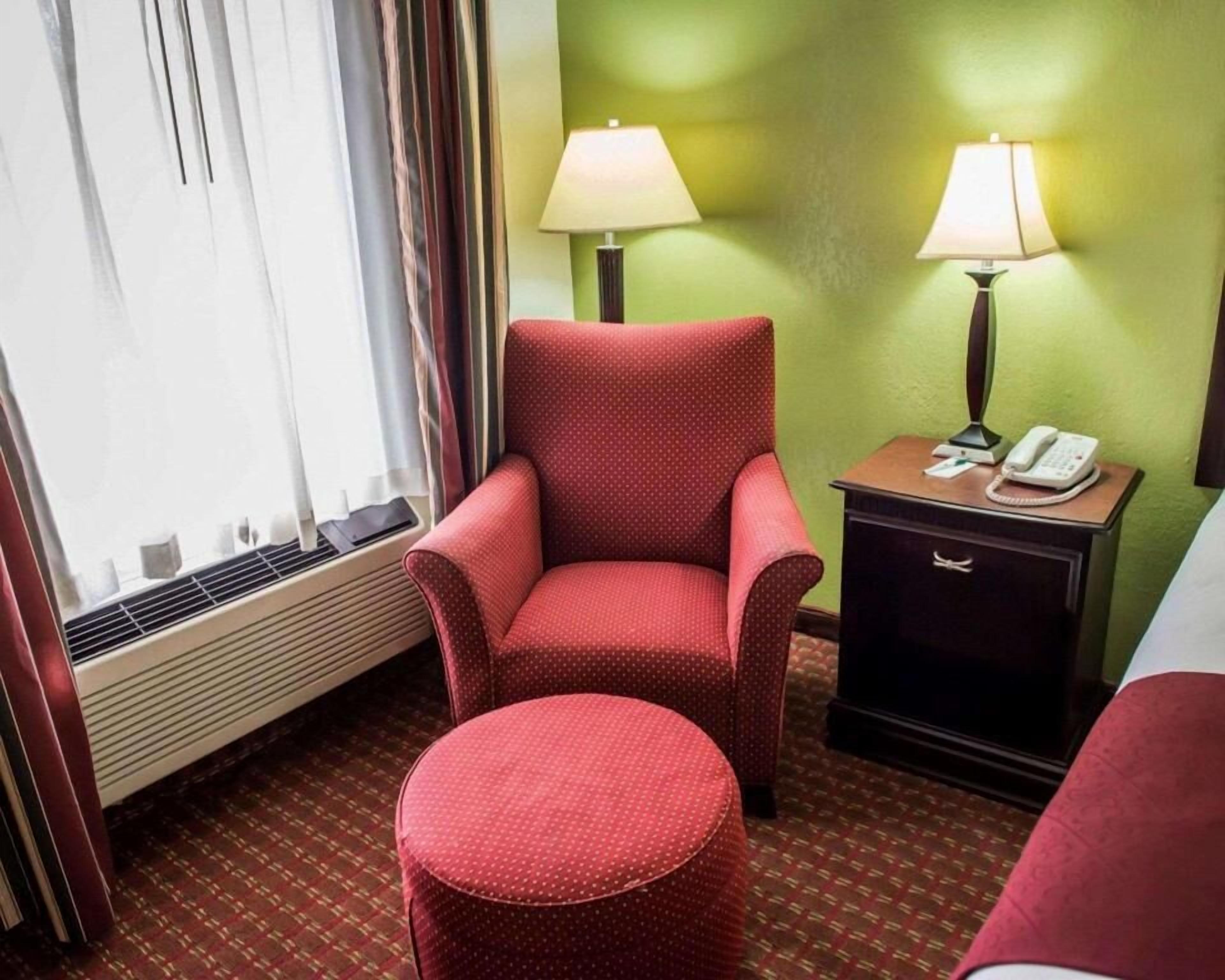 A hotel room with a red armchair, a small ottoman, a bedside table with a lamp, and a telephone.