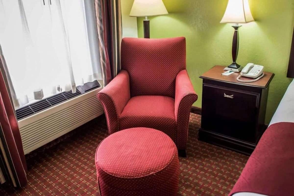 A hotel room with a red armchair, a small ottoman, a bedside table with a lamp, and a telephone.
