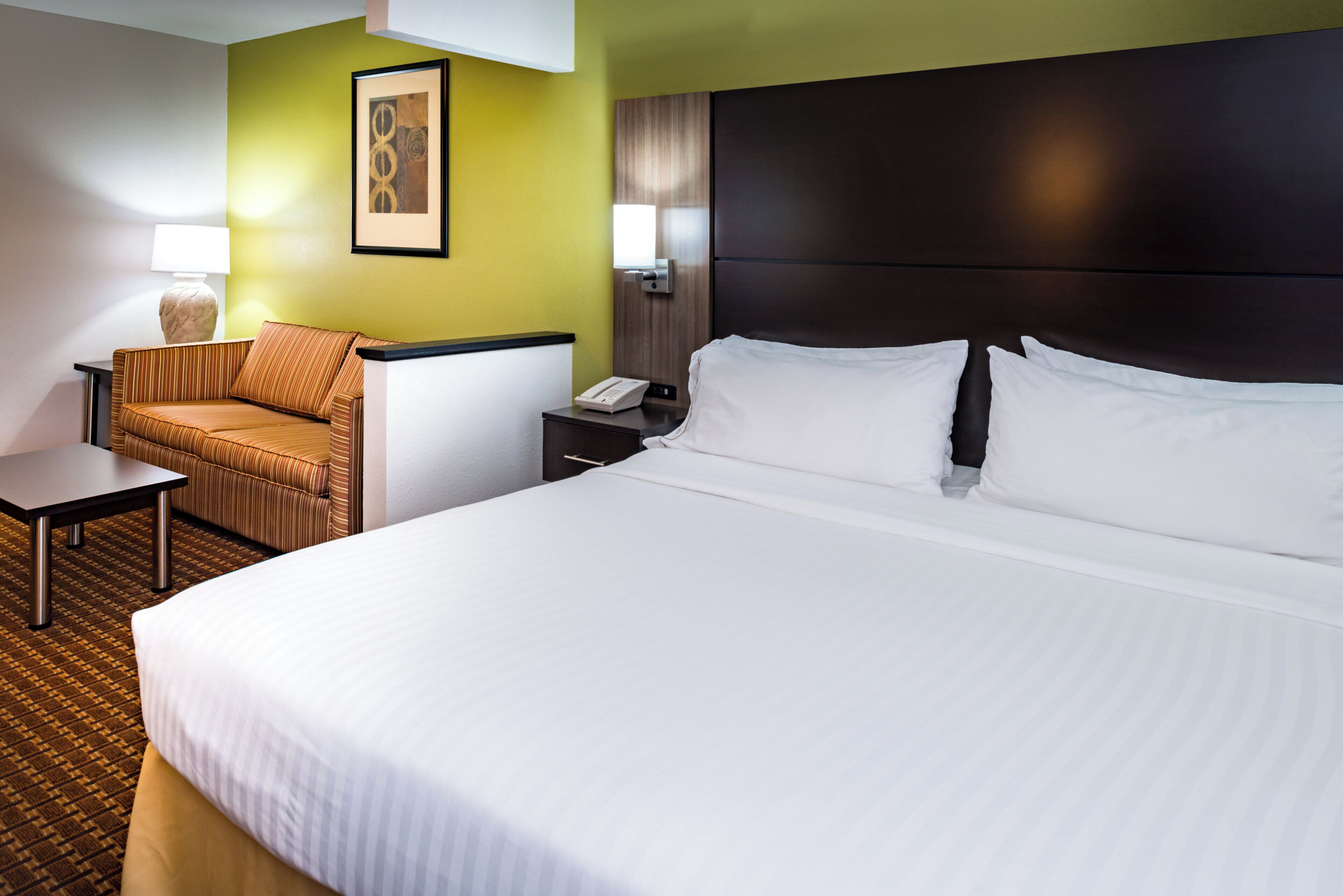 Habitación de hotel con una cama grande, una silla, una mesita pequeña, una mesita de noche y una lámpara.