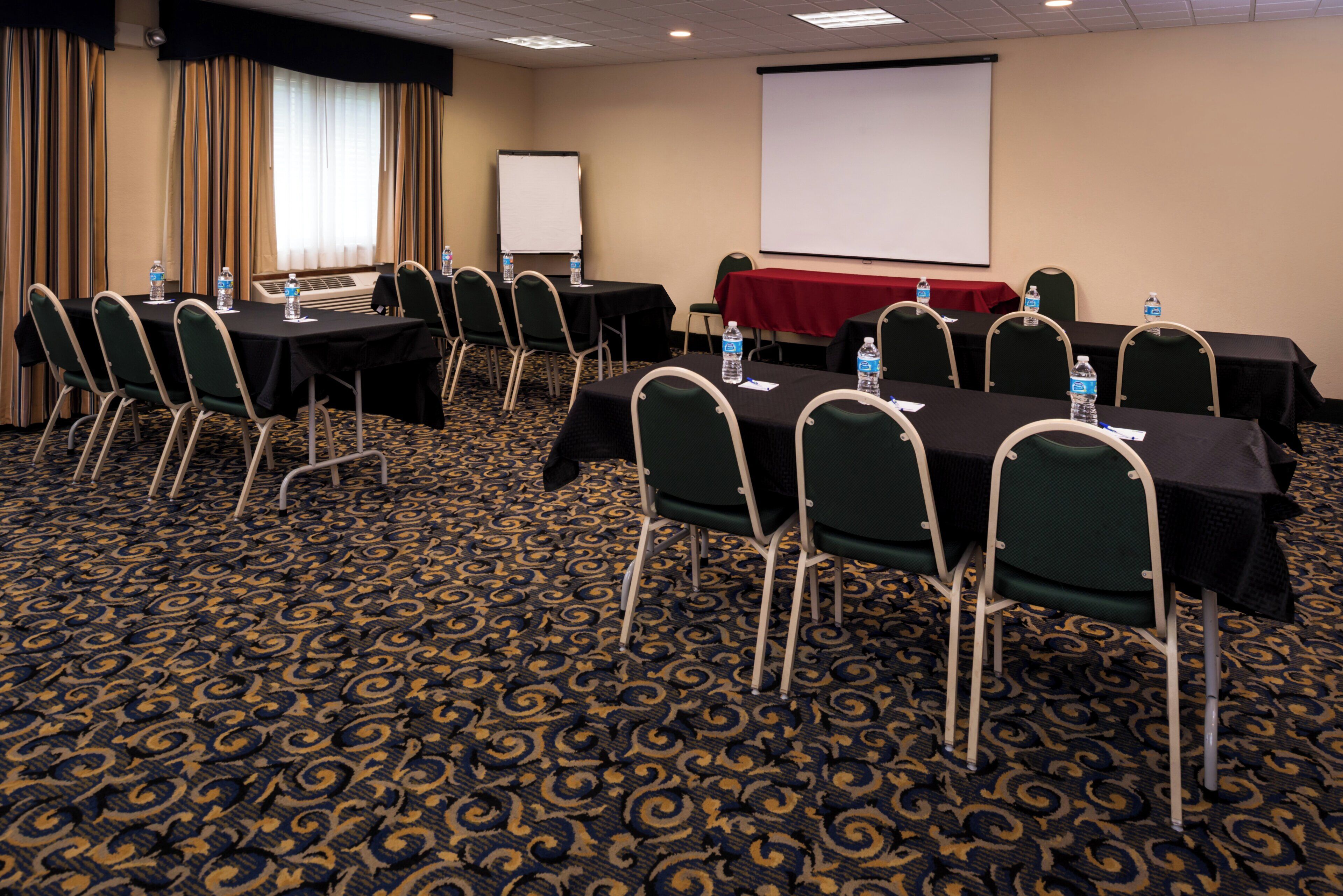 Un salón de conferencias con mesas, sillas, una pantalla para proyector y botellas de agua.
