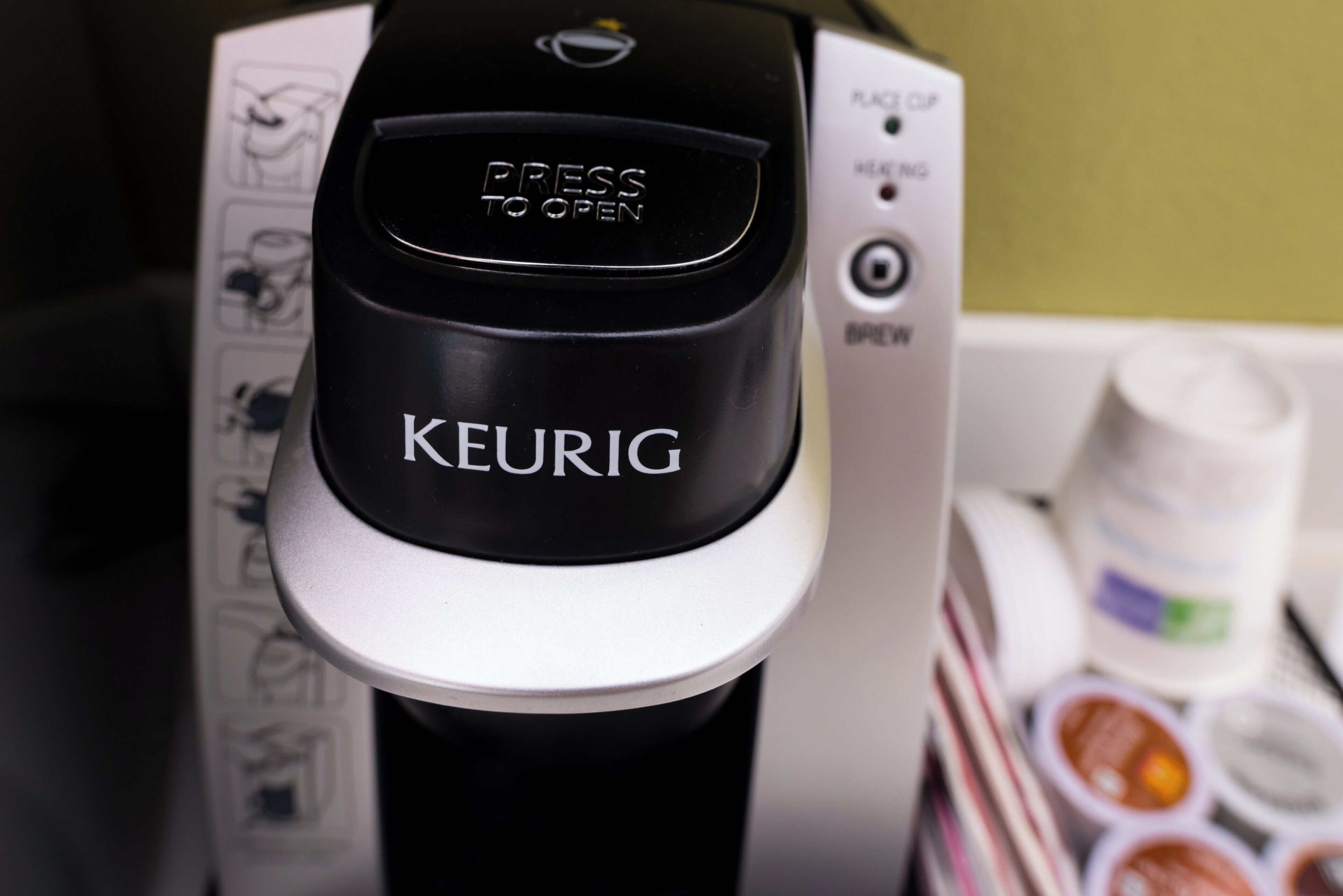 A Keurig coffee maker with a 'Press to Open' button, a 'RACE' indicator, and a 'BREW' button.