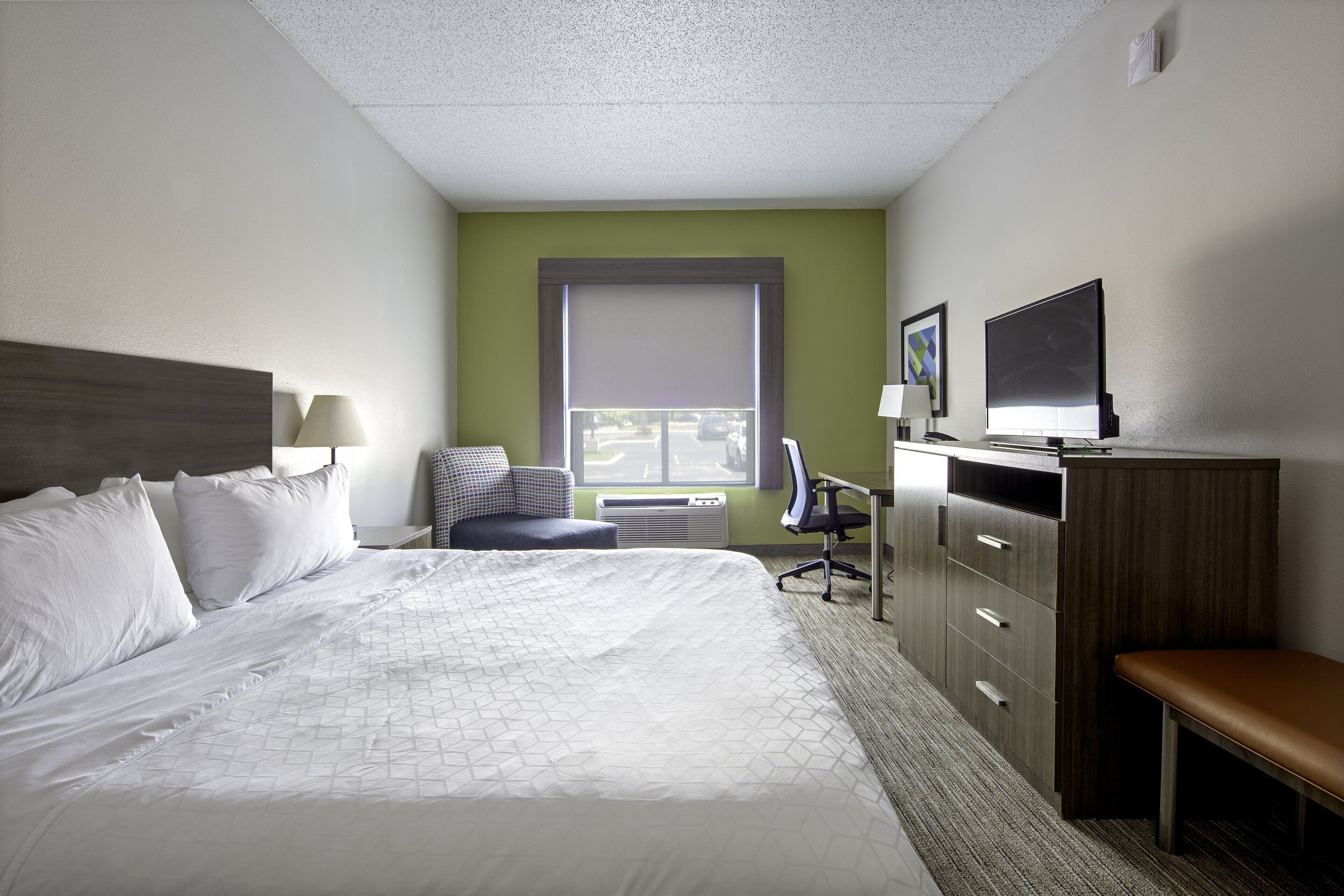 Quarto de hotel com uma cama grande, uma escrivaninha, uma cadeira, uma televisão e uma janela com persianas.