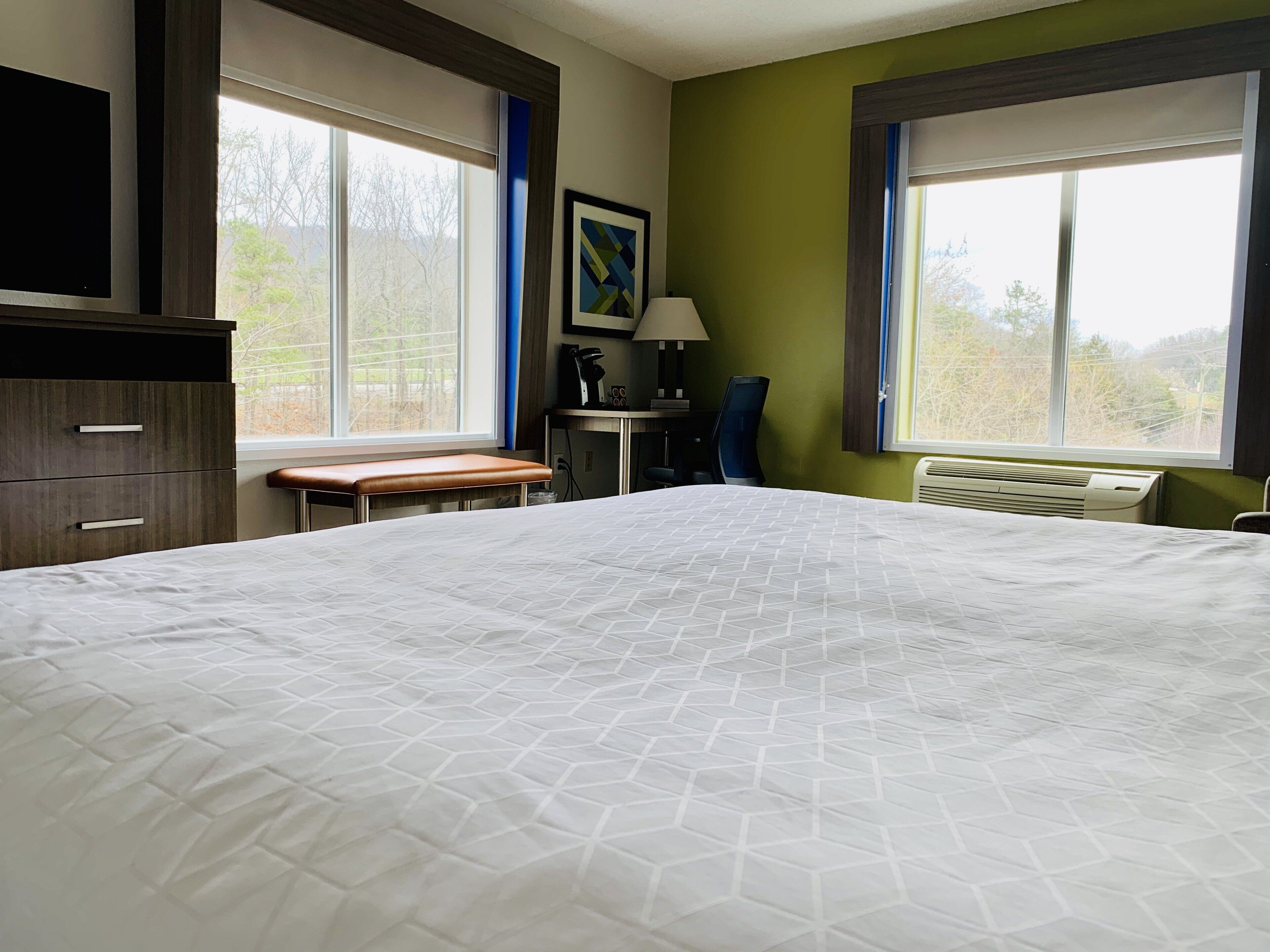 Habitación de hotel con una cama grande, un escritorio con silla, un banco y una ventana con vista a árboles.