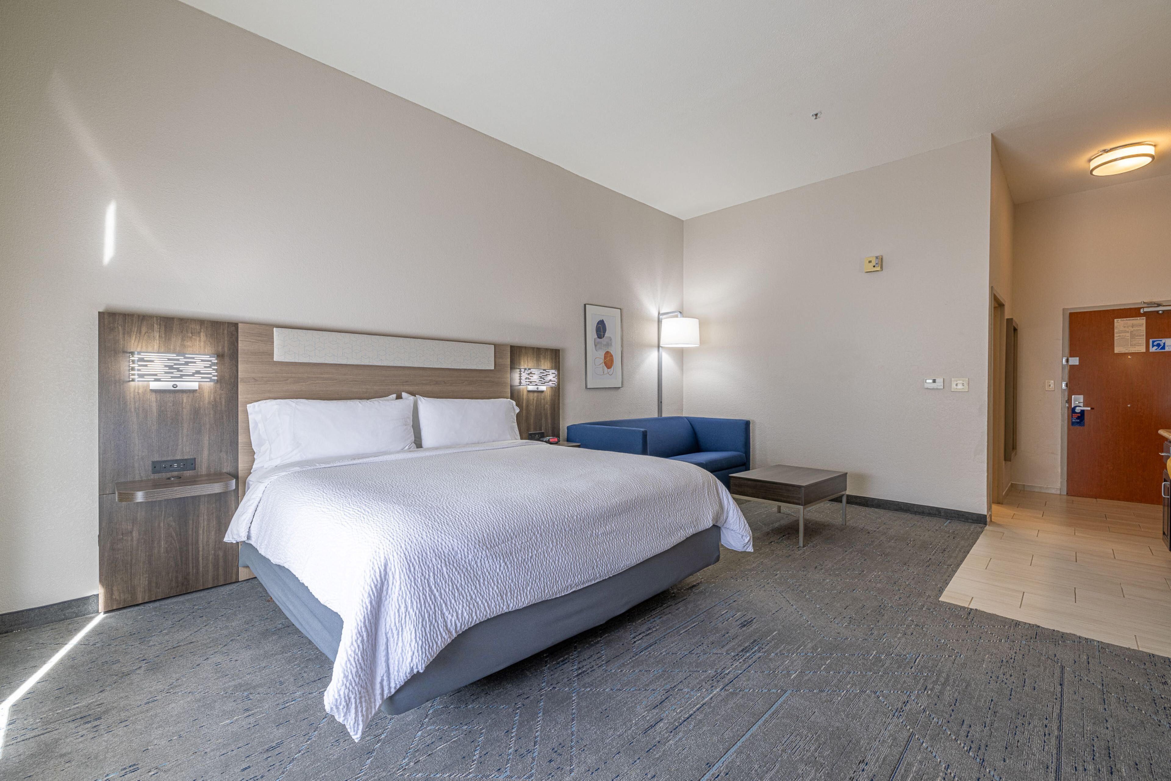 A modern hotel room with a large bed, a desk, a chair, and a wall-mounted lamp.