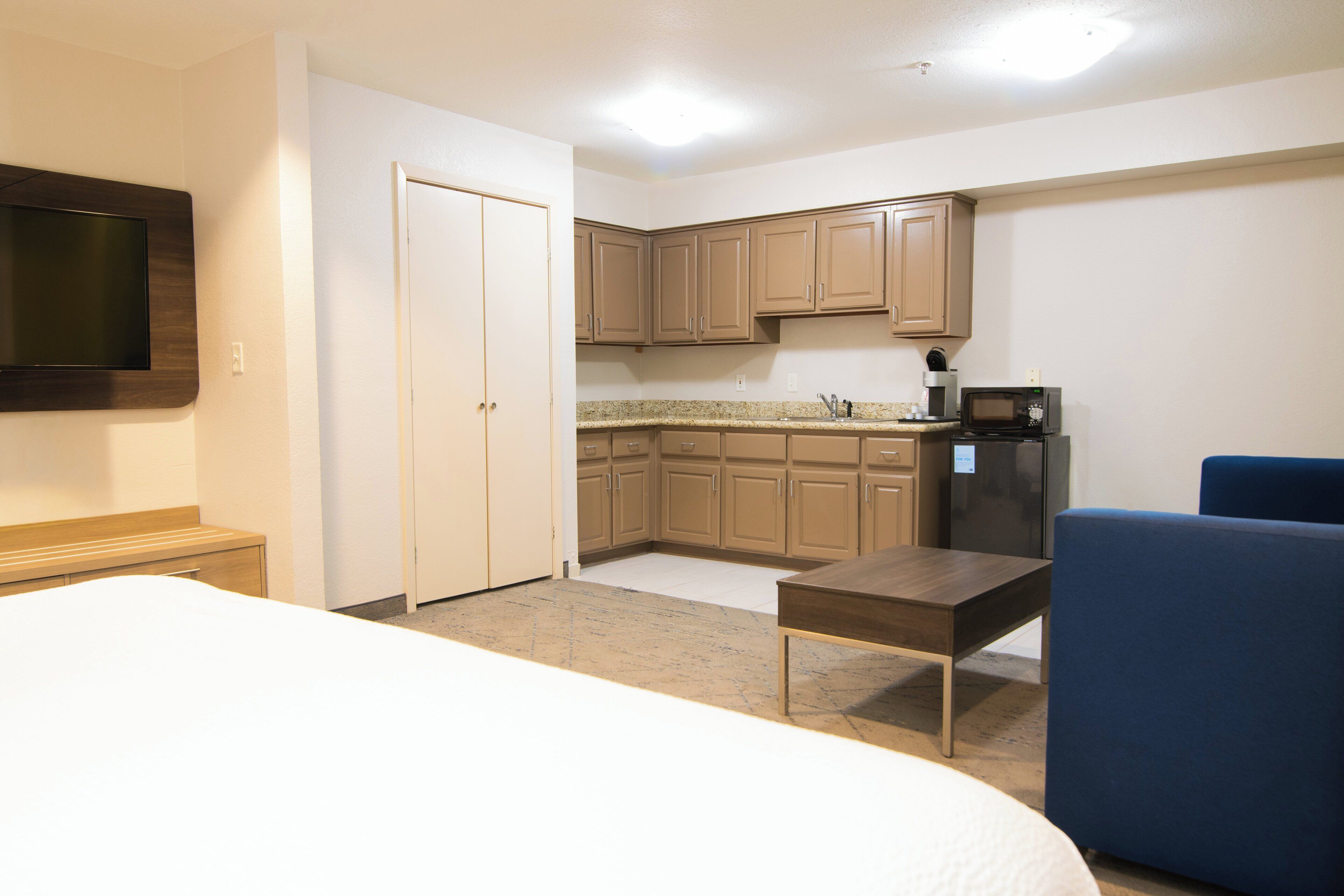 Una habitación de hotel con cama, escritorio, silla, televisión, kitchenette y puerta.