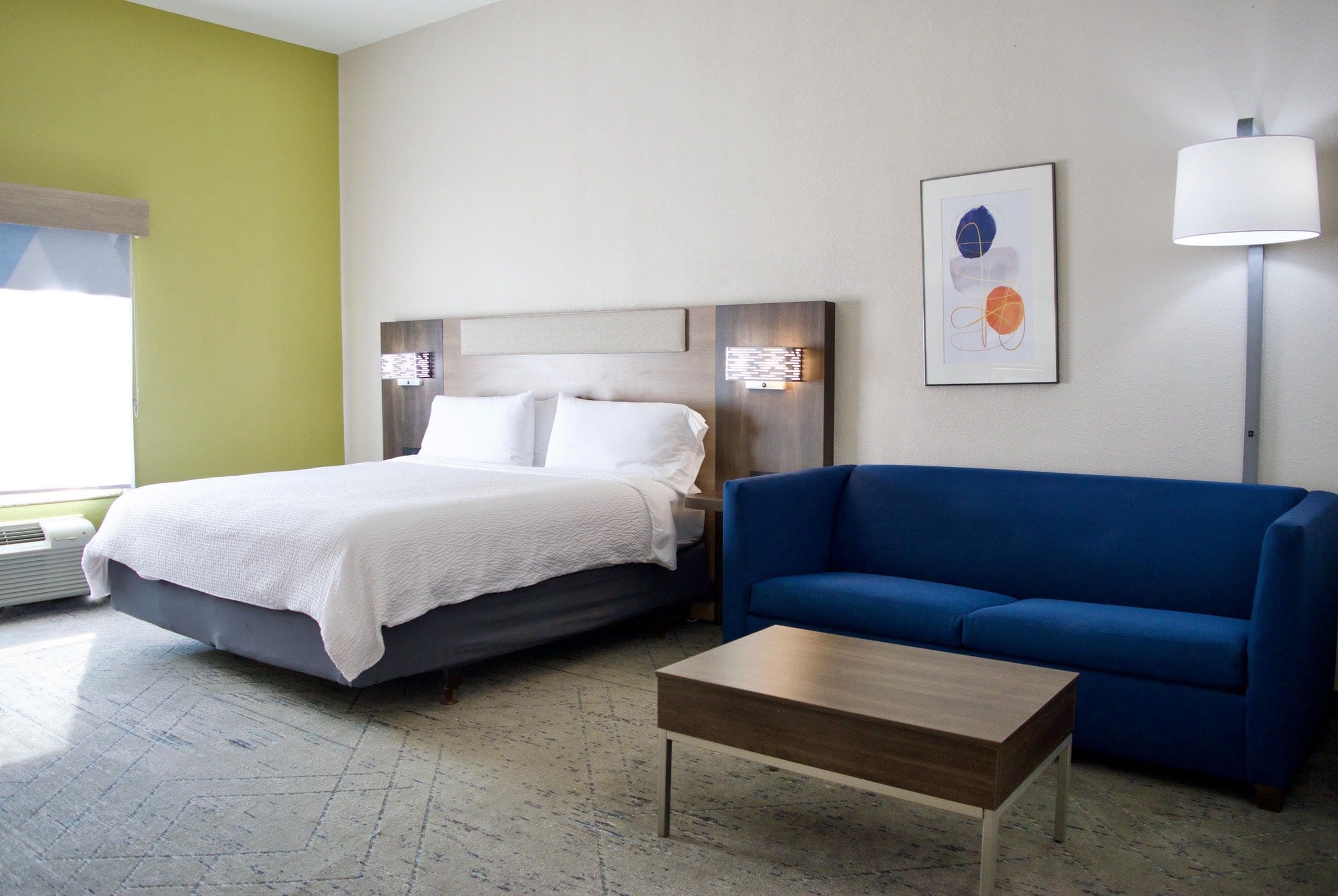 Una habitación de hotel con cama, sofá azul, mesita de madera y lámpara de pared.