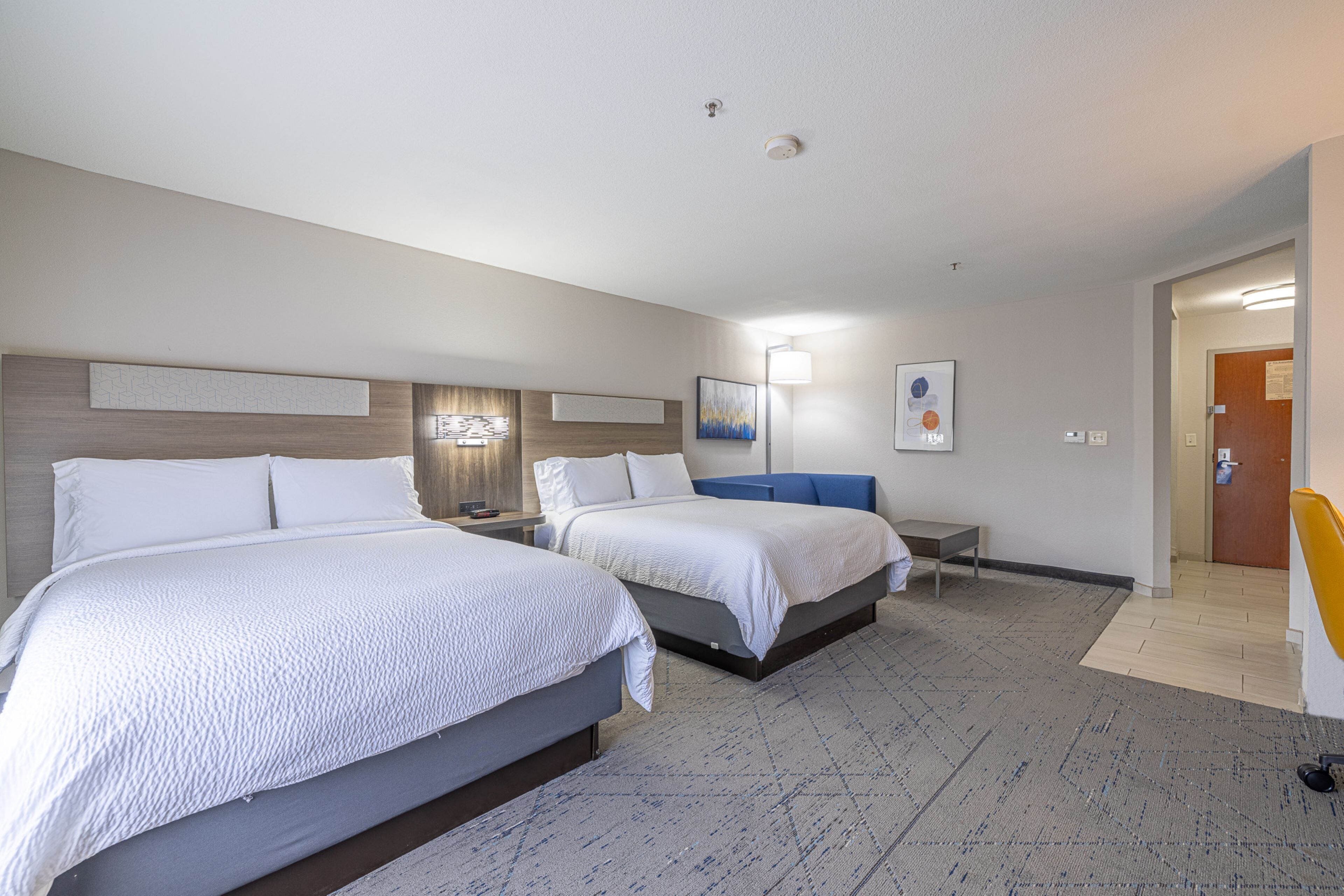 Habitación de hotel con dos camas, una mesita de noche con lámpara, un escritorio pequeño y una puerta que da a otra habitación.