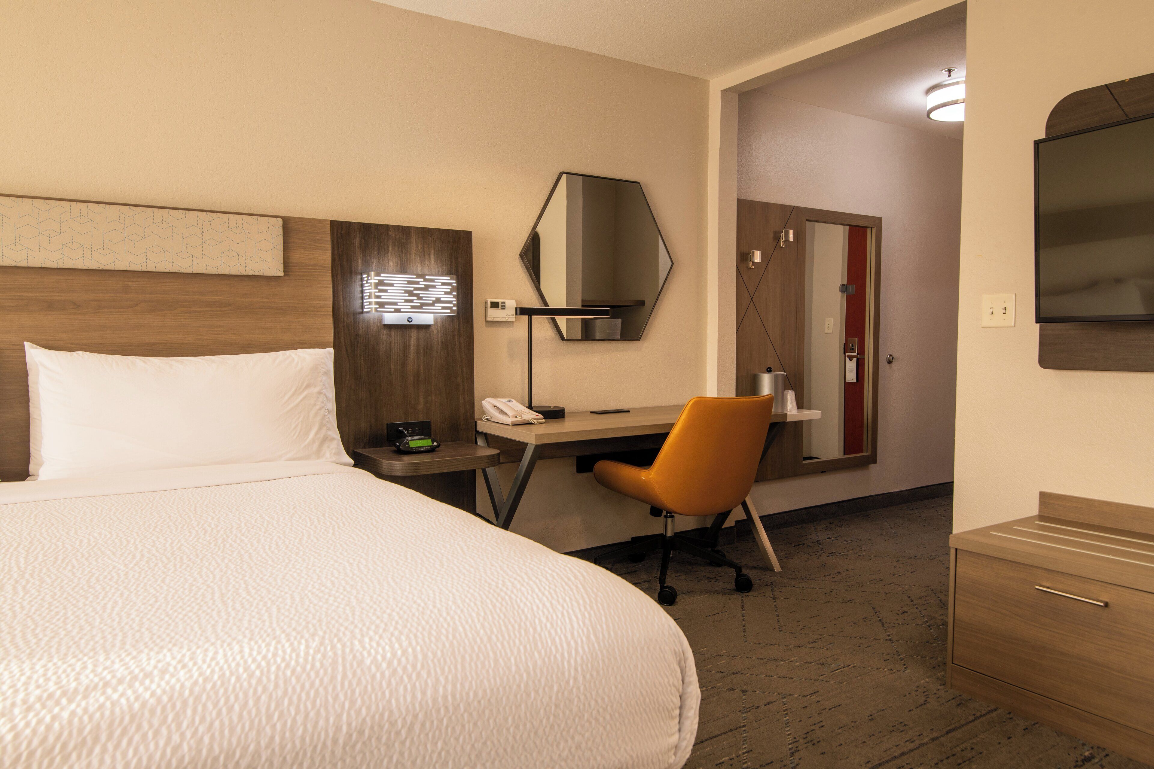 A hotel room with a large bed, a desk with a chair, a mirror, a TV, and a wardrobe.
