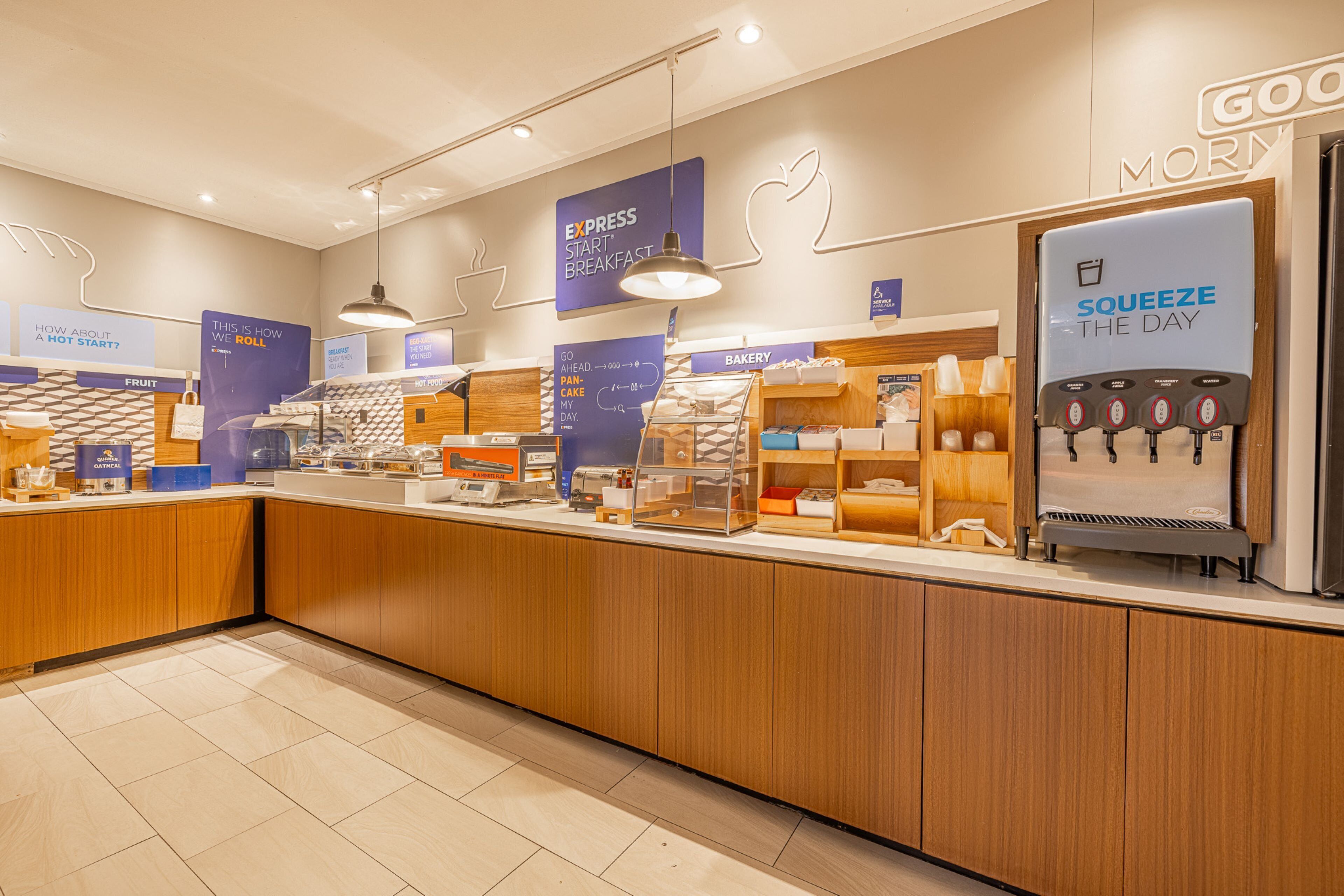 Un espace réservé au petit-déjeuner, avec un comptoir où sont exposés divers aliments, un distributeur d’eau et des panneaux indiquant les services proposés, tels que « PETIT-DÉJEUNER EXPRESS » et « PASSEZ UNE EXCELLENTE JOURNÉE À DÉGUSTER DES CRÊPES ».