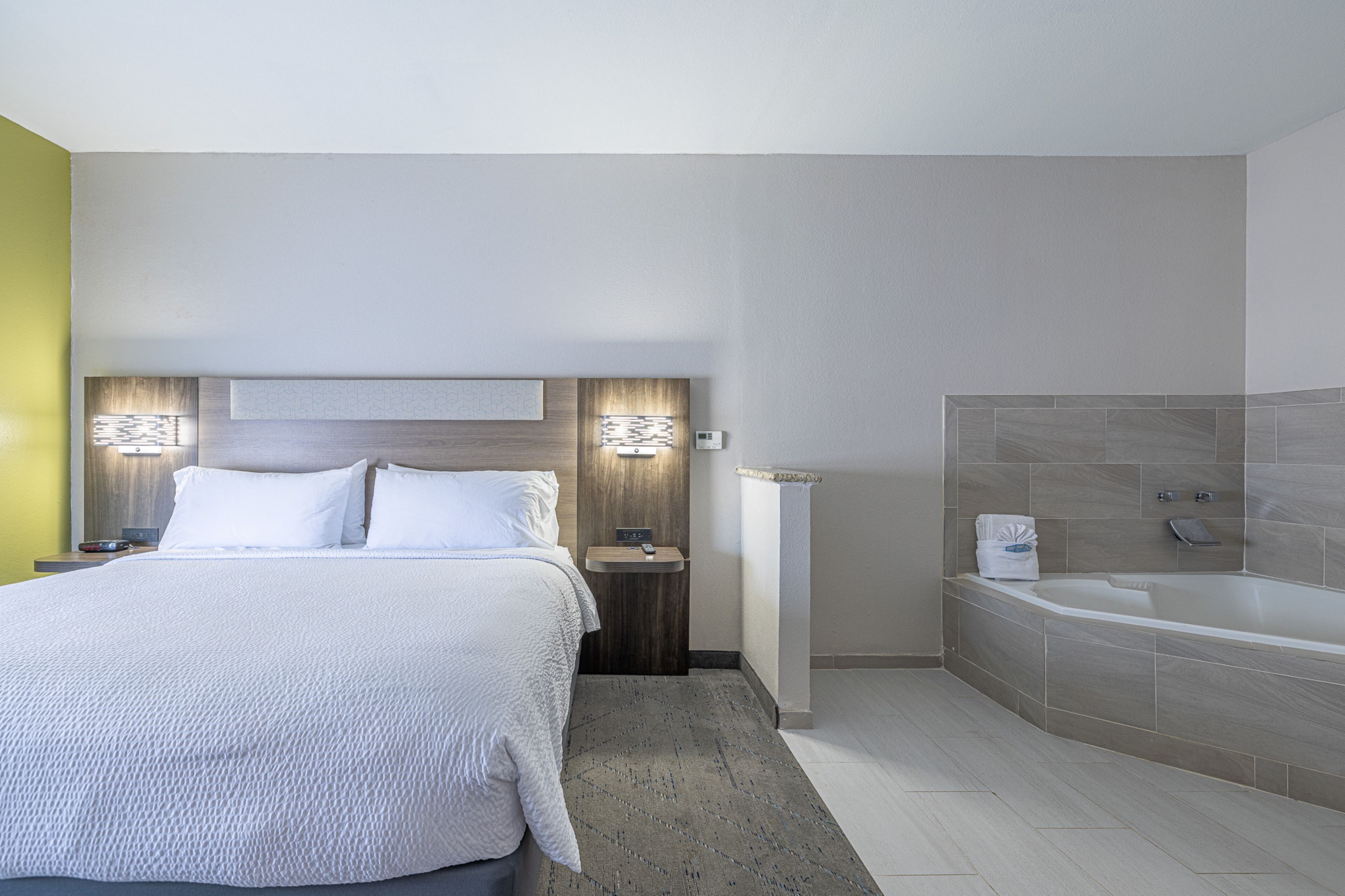 Una habitación de hotel moderna con una cama grande, mesitas de noche, una lámpara de pared y un baño con bañera y ducha.