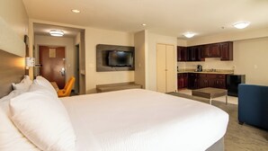A hotel room with a large bed, a television mounted on the wall, a small kitchenette, and a chair.