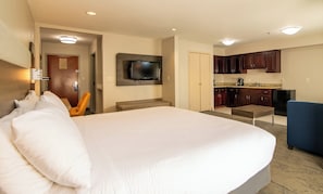 Una habitación de hotel con una cama grande, televisión instalada en la pared, una kitchenette pequeña y una silla.