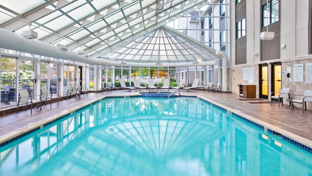 An indoor swimming pool with a glass ceiling, surrounded by lounge chairs and a view of the outdoors.