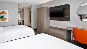 A hotel room with two beds, a TV mounted on the wall, a desk with a chair, and a wardrobe.