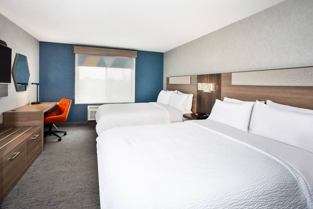 Una habitación de hotel con dos camas, un escritorio, una silla naranja y un ventanal grande.