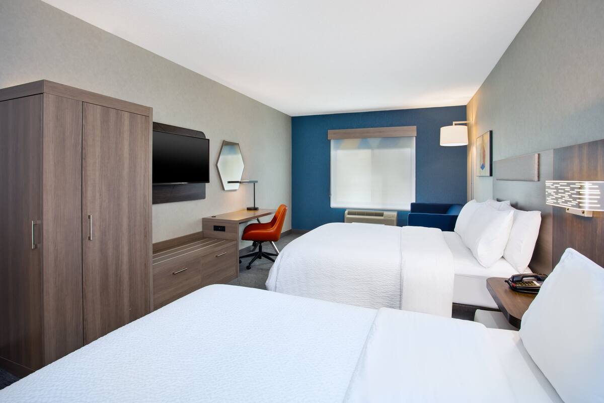 Habitación de hotel con dos camas, un escritorio con silla, televisión y ventana con persianas.