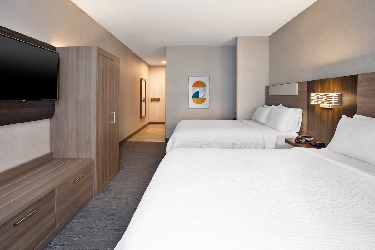 Habitación de hotel con una cama grande, un televisor montado en la pared, un armario de madera y una mesita de noche con una lámpara.