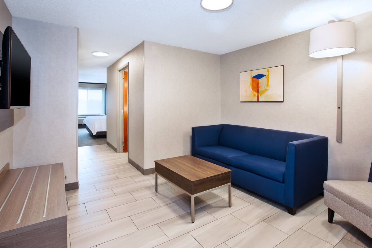 A modern hotel room with a wooden coffee table, a blue sofa, a flat-screen TV mounted on the wall, and a painting above the sofa.