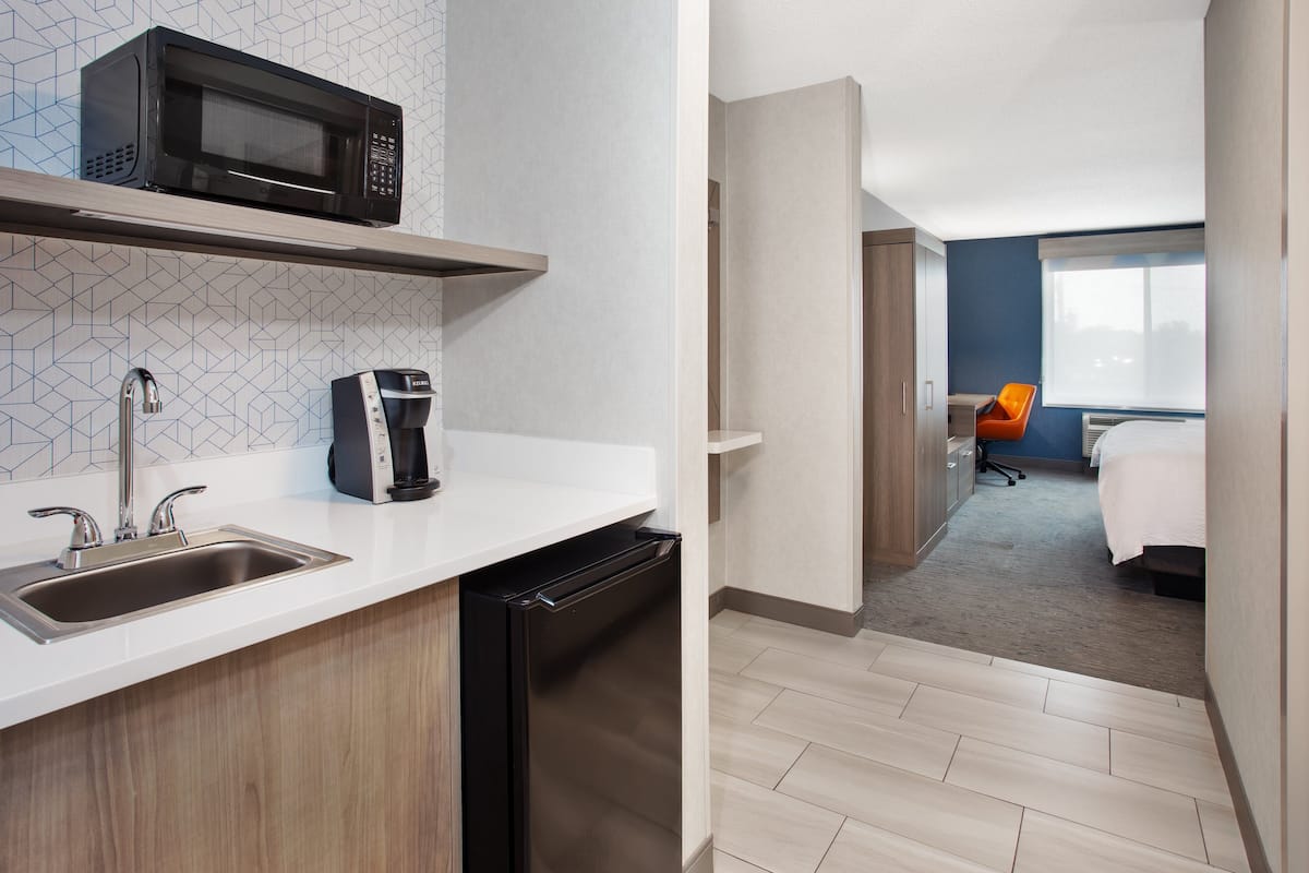 Una habitación de hotel moderna con un área de cocina equipada con microondas, cafetera y lavavajillas. La habitación tiene una cama, un escritorio y una ventana con vistas.