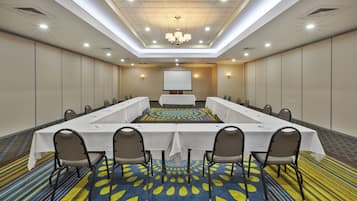 Un salón de conferencias con una mesa larga, sillas, una pantalla de proyector y una alfombra estampada.