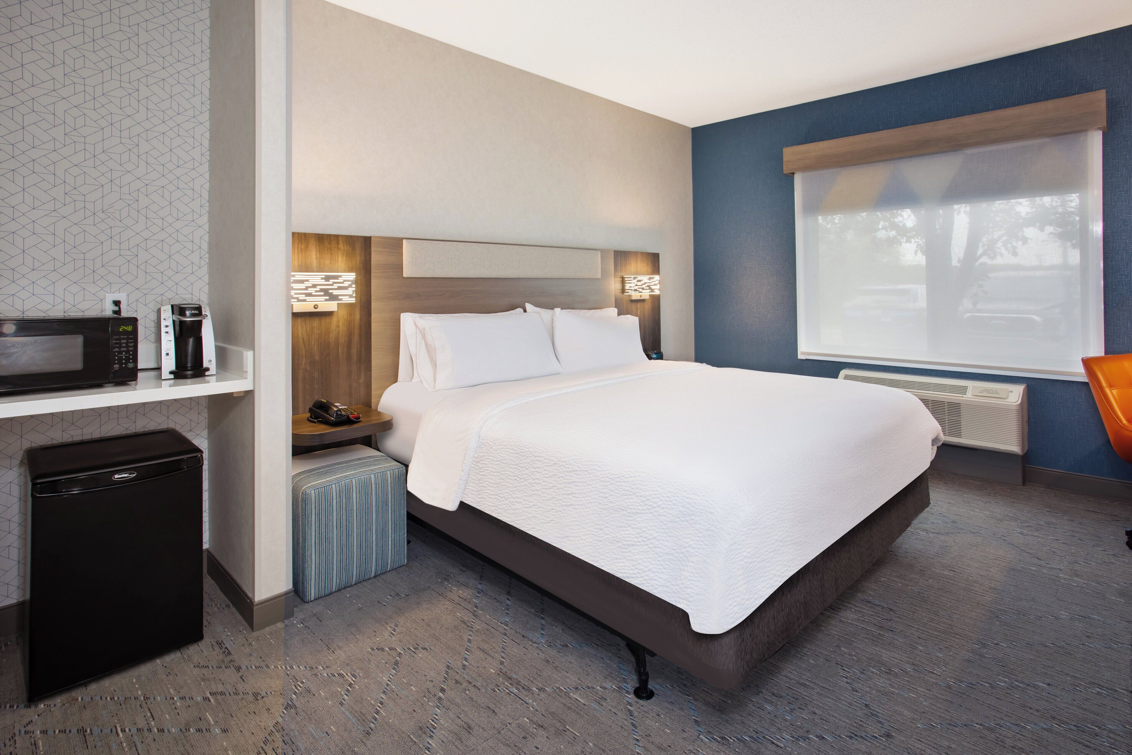 A hotel room with a bed, bedside table, lamp, and a view of the outdoors.