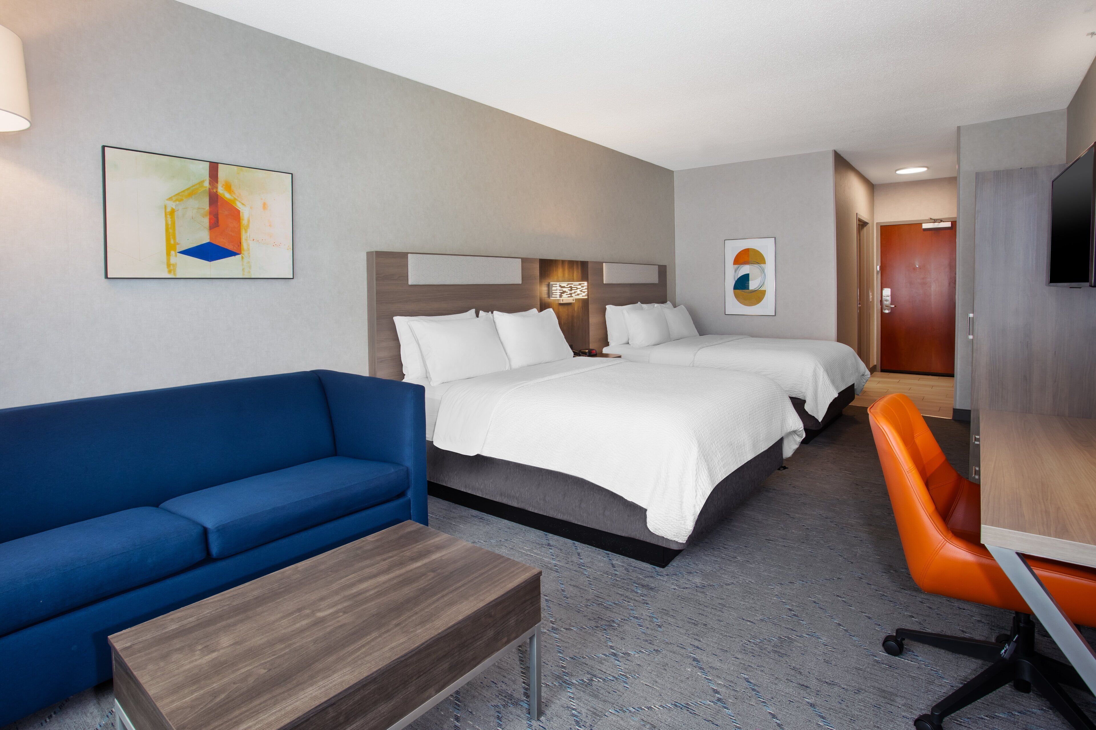 A hotel room with a bed, a blue sofa, an orange chair, a wooden table, and artwork on the walls.