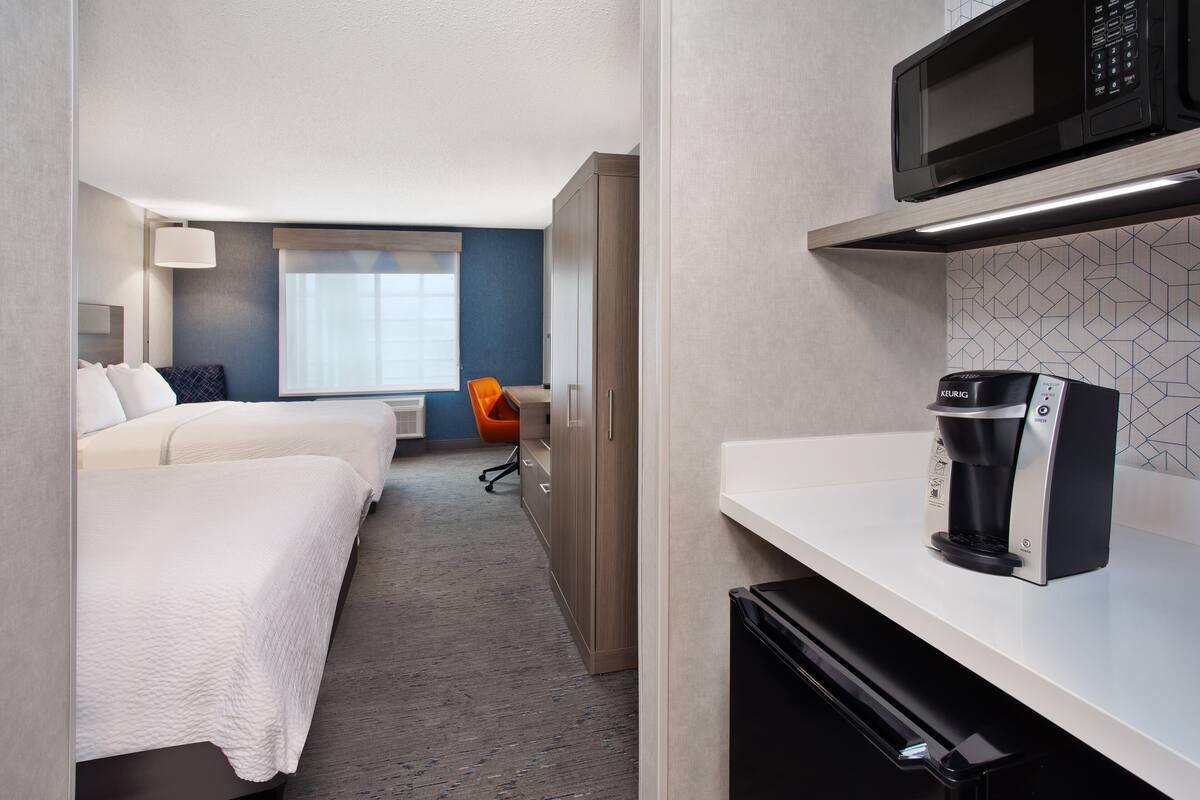 Habitación de hotel con kitchenette, cama con ropa blanca, una silla y ventana con persianas.