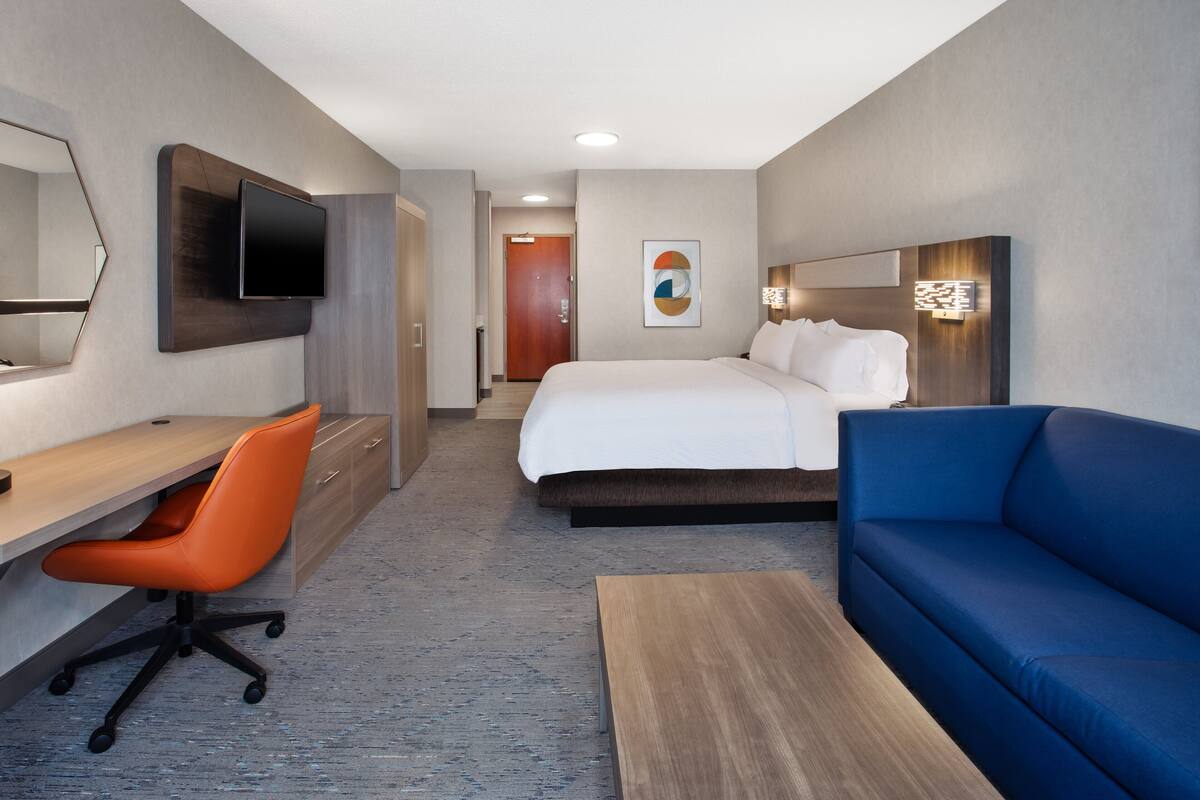 Una habitación de hotel con una cama, un escritorio con una silla naranja, un sofá azul, un televisor montado en la pared y un cuadro en la pared.