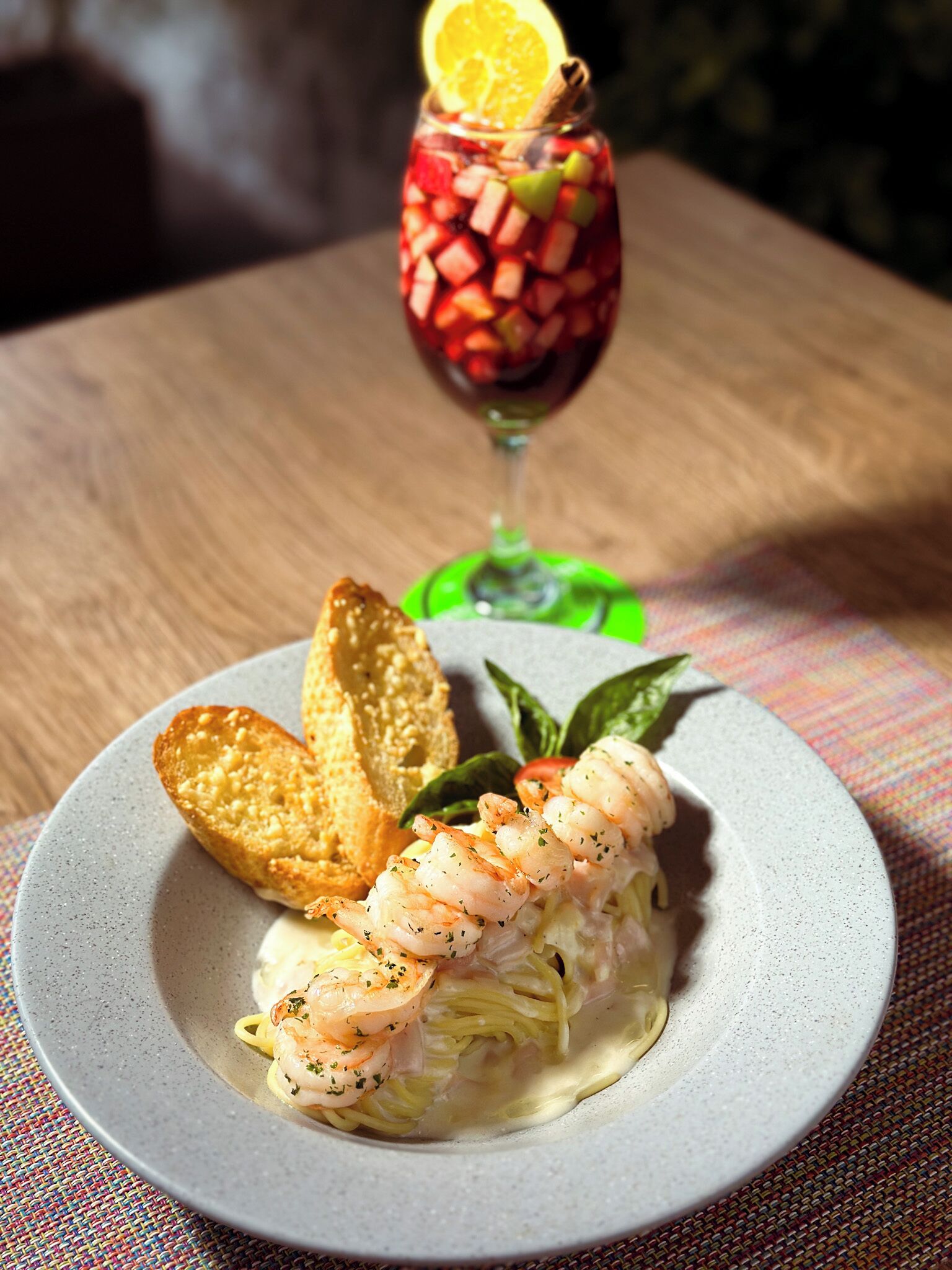 Un plato de pasta con mariscos y camarones, adornado con hierbas, acompañado de pan tostado y una copa de vino tinto con una rodaja de naranja.