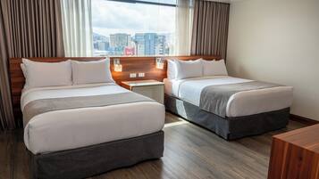 A hotel room with two beds, wooden headboards, a nightstand with a lamp, and a view of the cityscape.