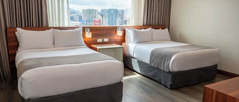 A hotel room with two beds, wooden headboards, a nightstand with a lamp, and a view of the cityscape.