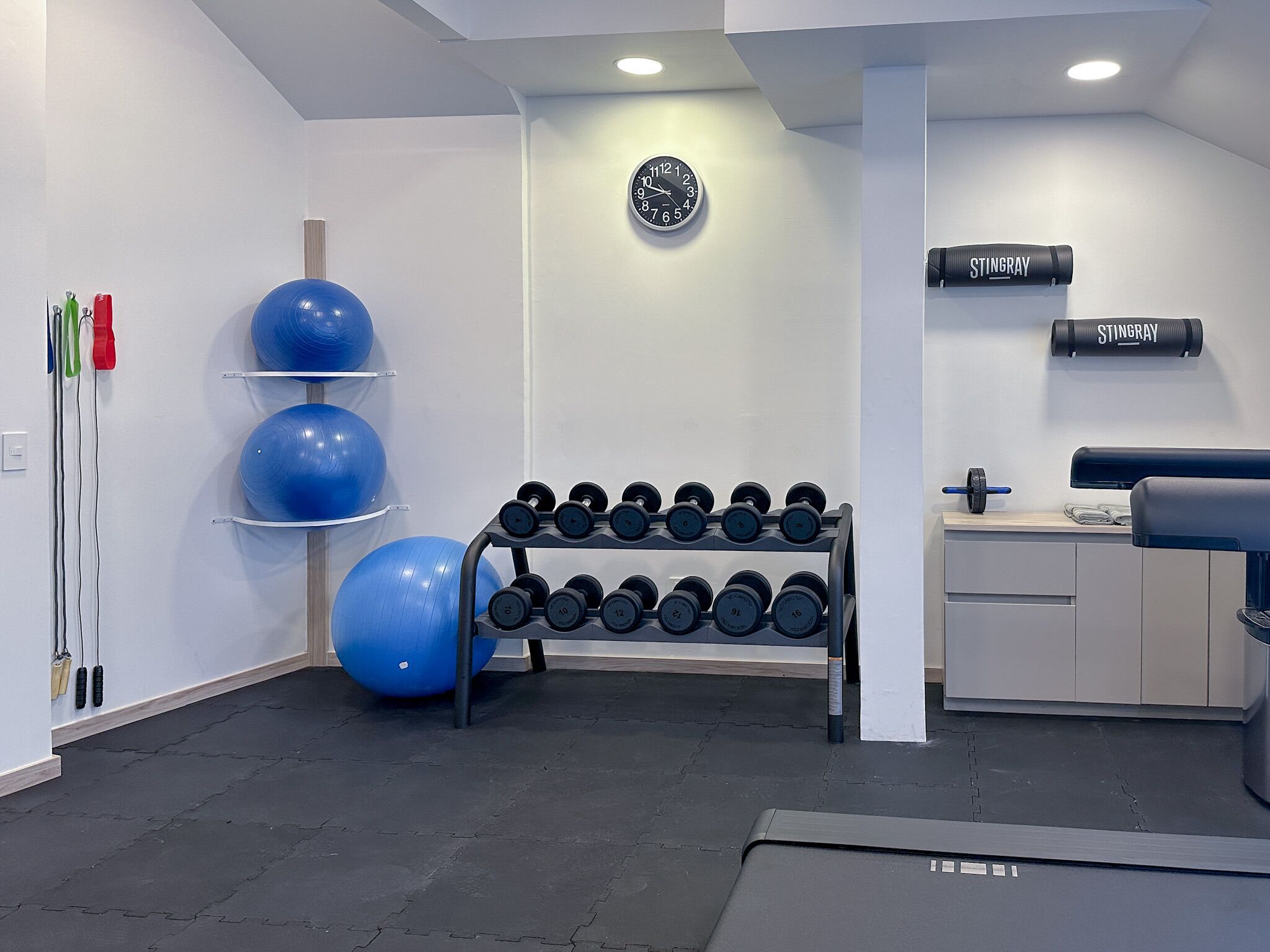 Un gimnasio con equipamiento de ejercicio, incluyendo pesas y pelotas de gimnasia, y un reloj de pared.