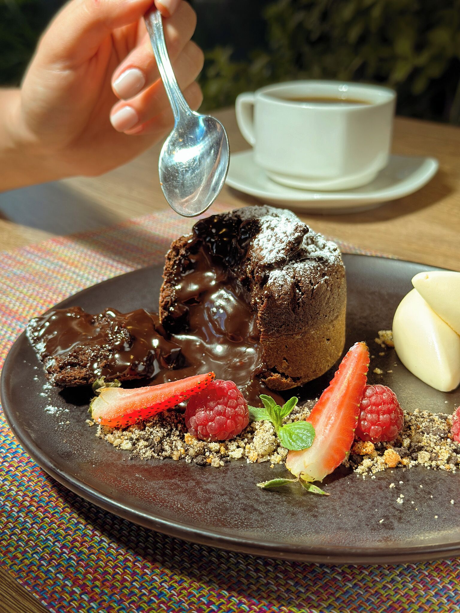 Un plato con postre que incluye un pastel volcán de chocolate, fresas, frambuesas y una hoja de menta, acompañado de una taza de café.