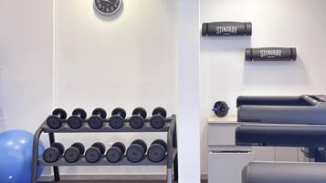 Un gimnasio con un reloj de pared, mancuernas, una pelota de ejercicios azul y una mesa de masajes.