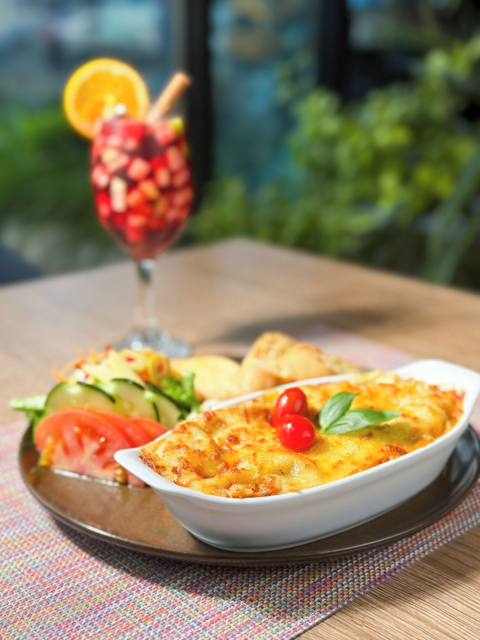 A meal with a baked dish, salad, and a drink on a table with a view of greenery.