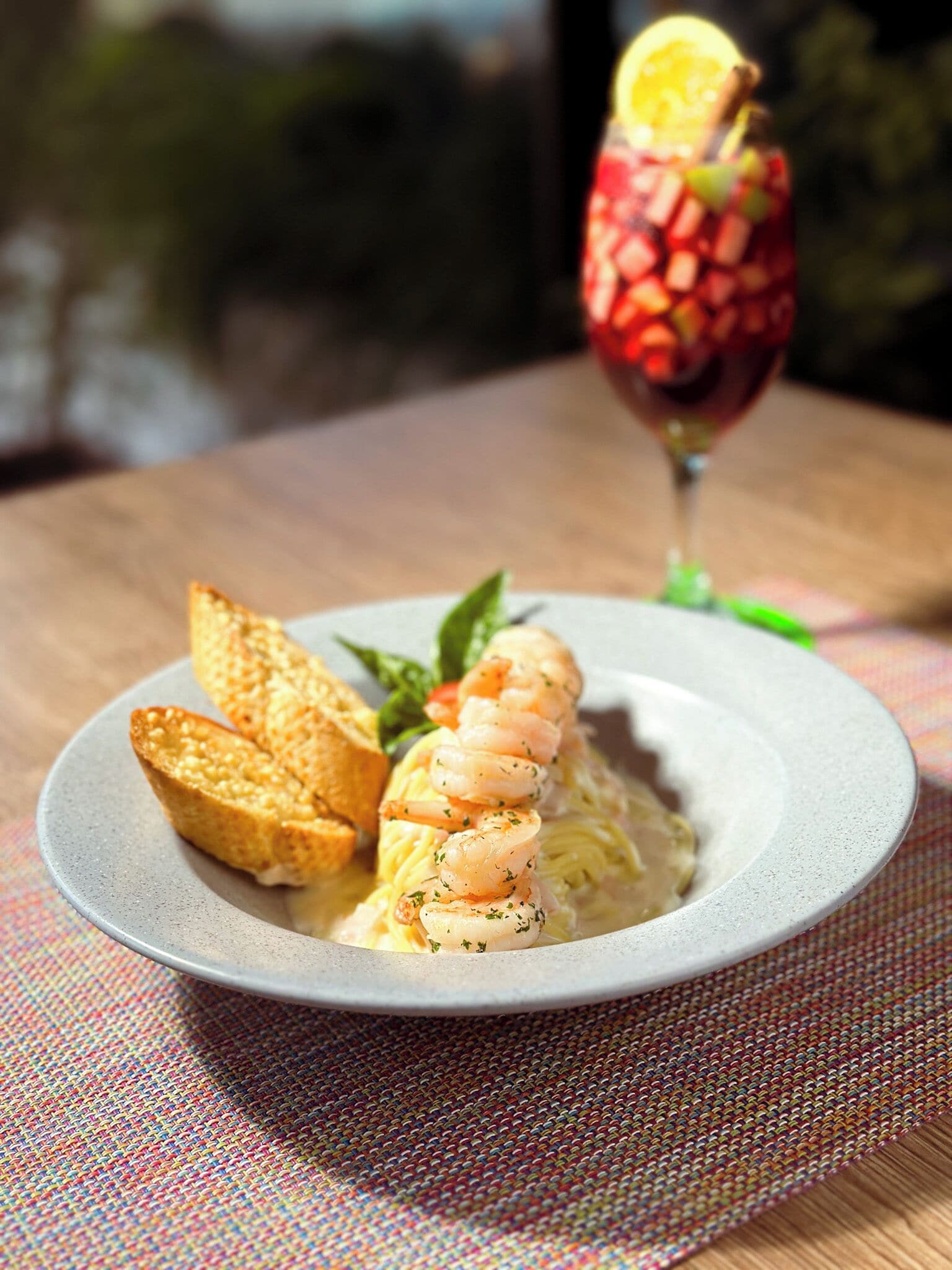 Un plato de pasta con camarones, un vaso de bebida roja y pan tostado sobre una mesa con un posavasos tejido.