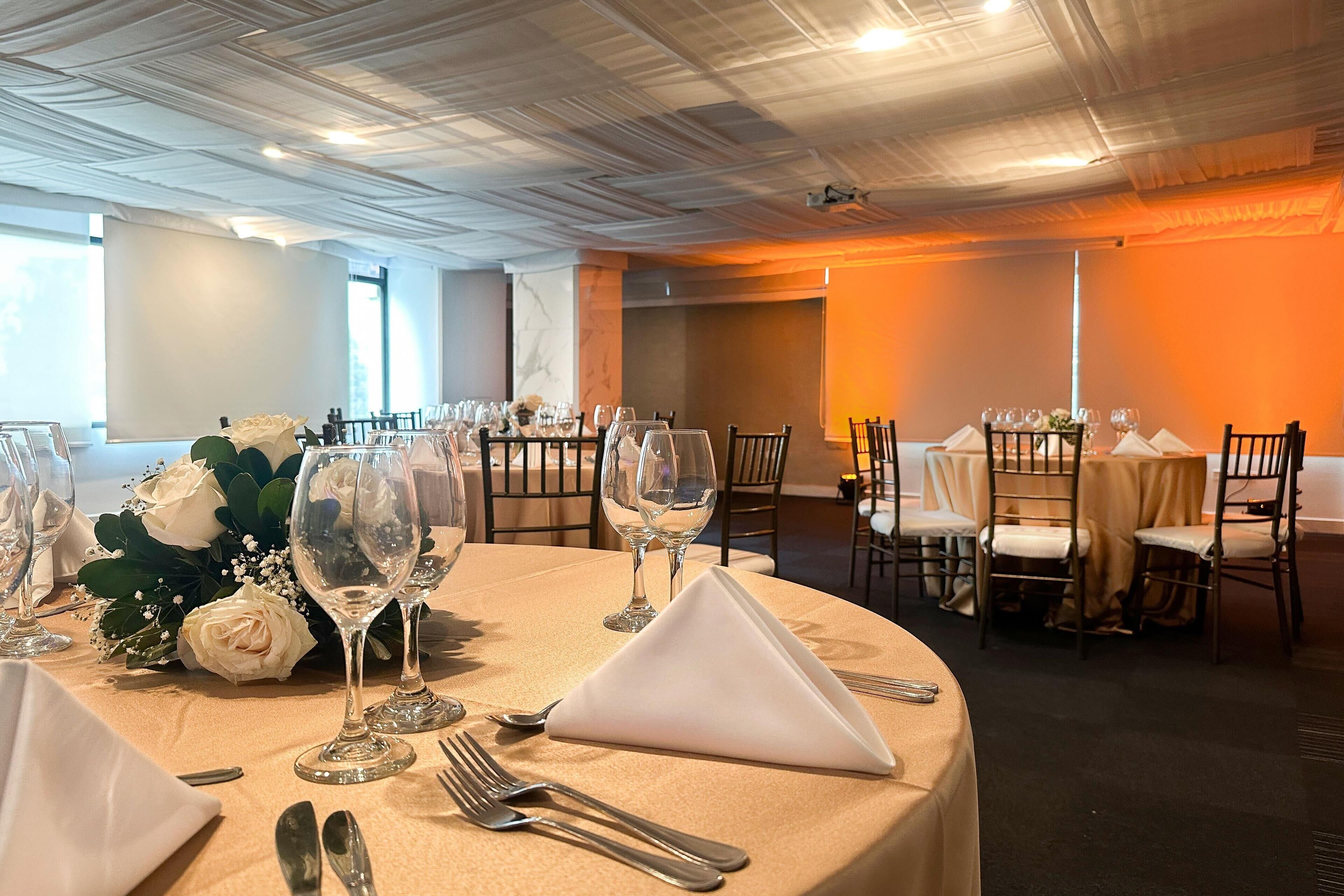 Un salón de banquetes con mesas redondas dispuestas para un evento formal, con manteles blancos, cubiertos y centros de mesa florales.