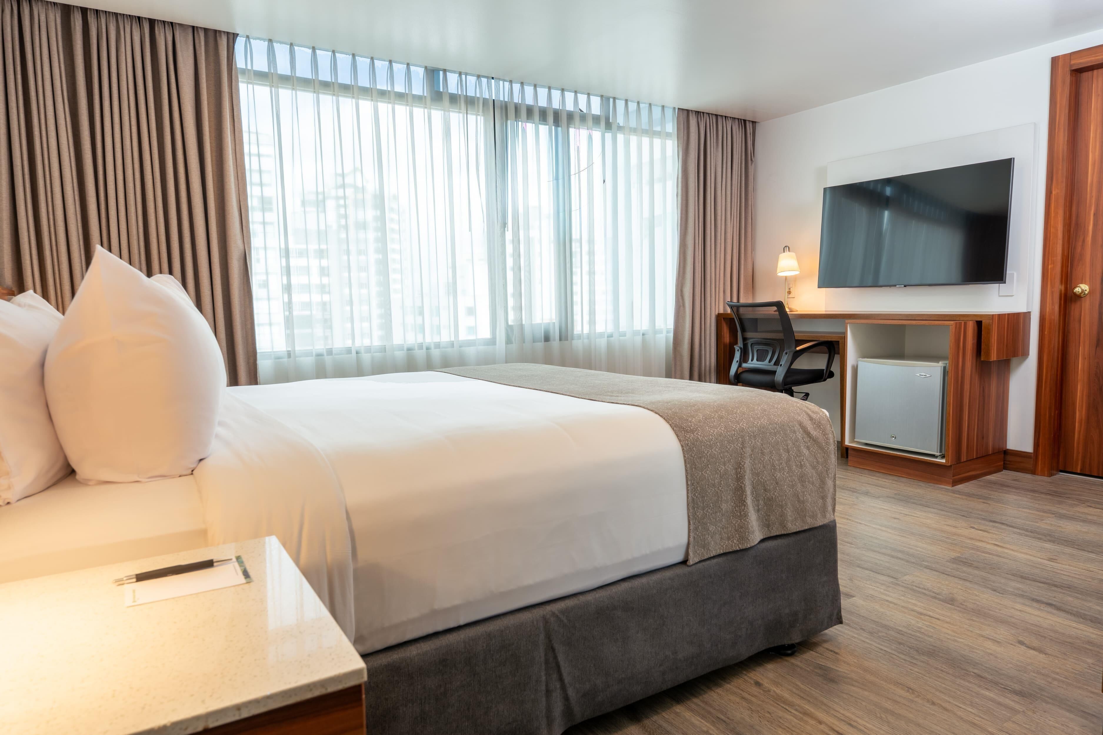 A hotel room with a large bed, a desk with a chair, a television, and a minibar.