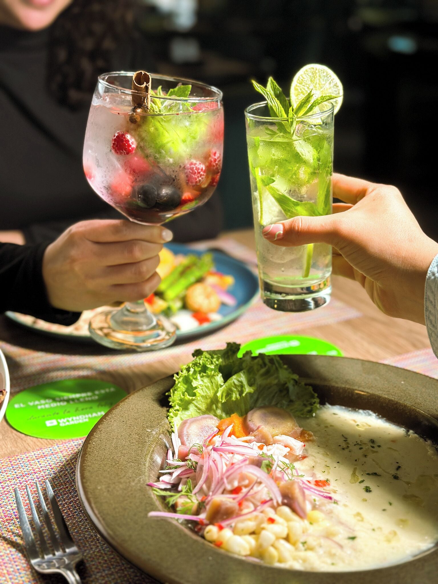 A dining table with a plate of ceviche, a glass of cocktail with lime and mint, and a fork on a colorful placemat.