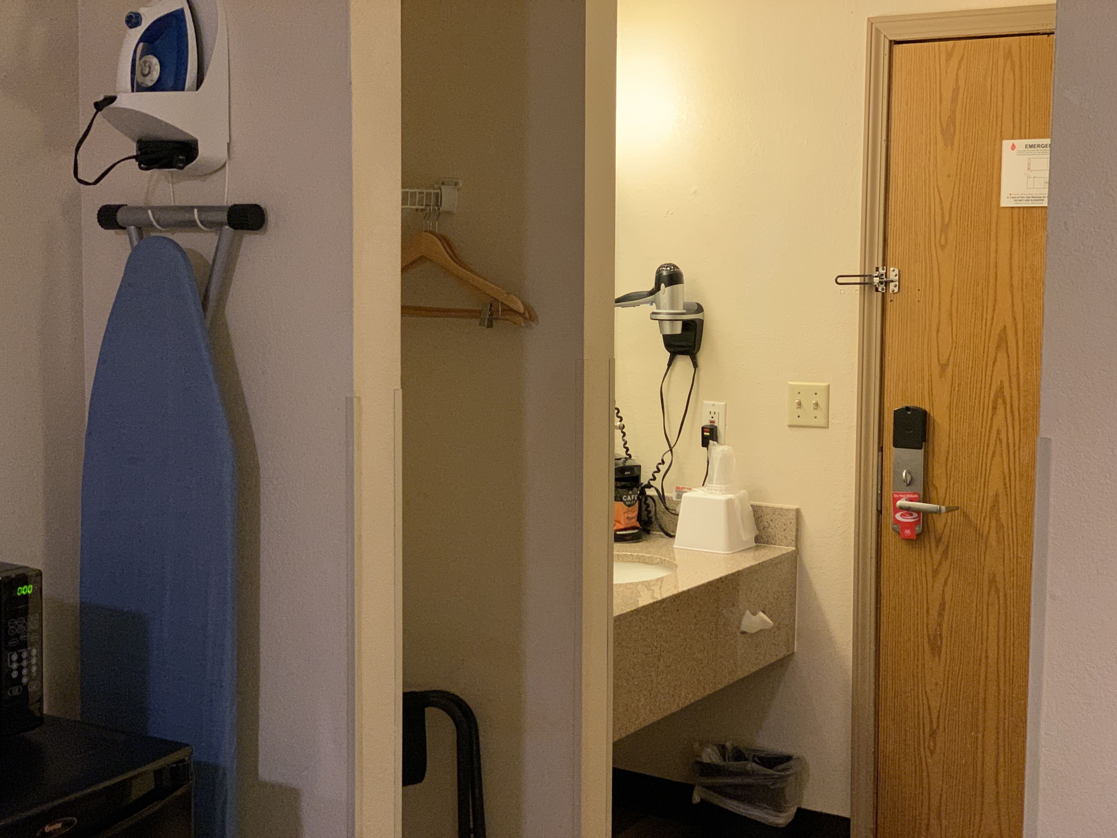 A hotel room with a wooden door, a microwave, an ironing board, a hairdryer, and a small trash bin.