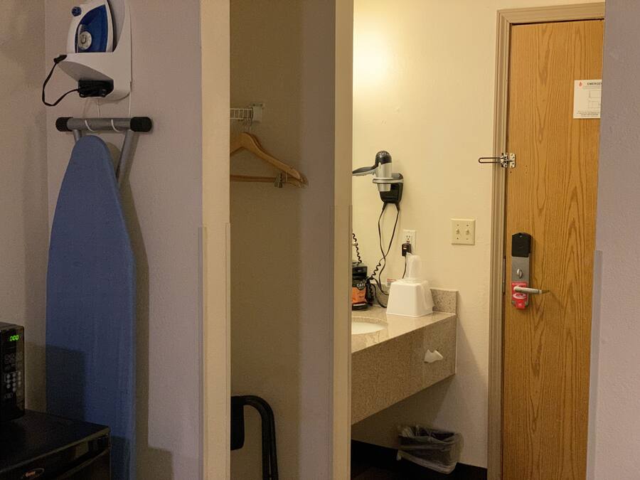 A hotel room with a wooden door, a microwave, an ironing board, a hairdryer, and a small trash bin.