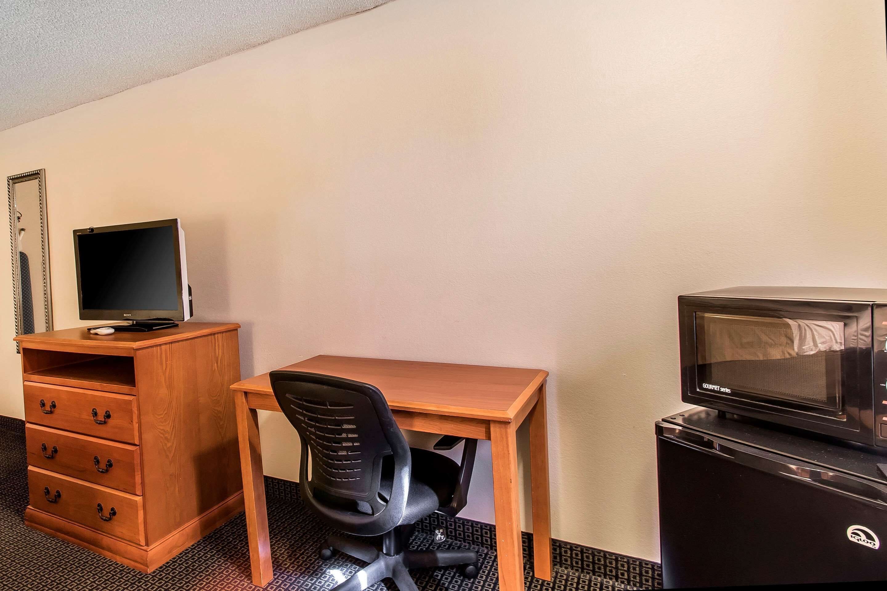Une petite chambre d’hôtel avec un bureau en bois, une chaise de bureau noire, un téléviseur posé sur une commode en bois et un four à micro-ondes sur un réfrigérateur noir.